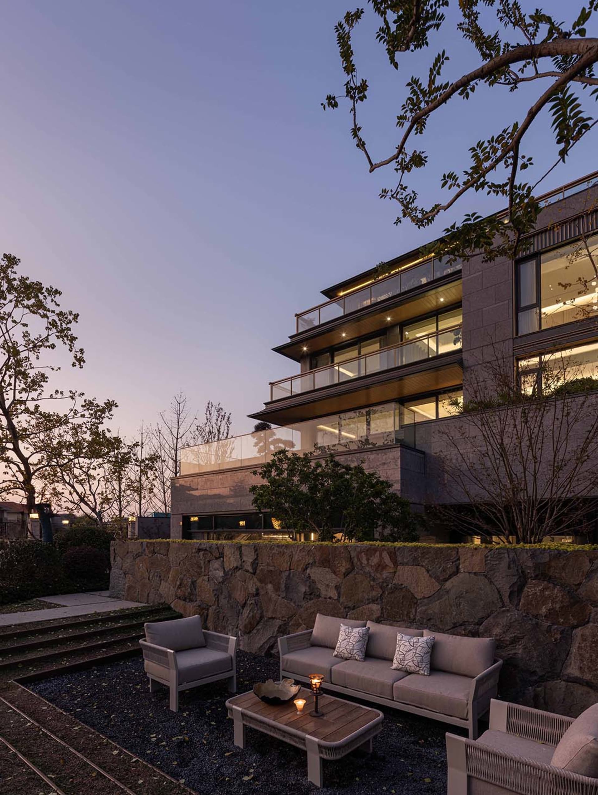 A modern building with a stone wall and patio area, featuring a glass wall, two couches, and a chair, creating a comfortable outdoor seating area with natural light and a warm atmosphere