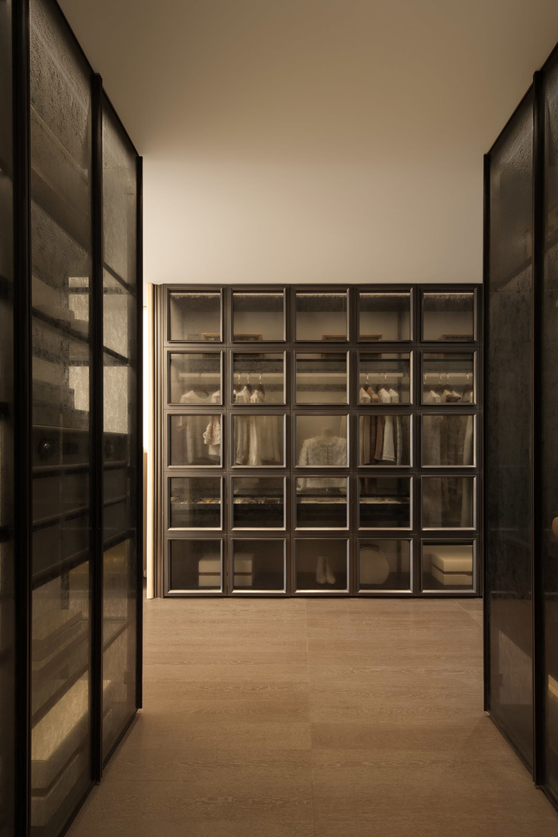 A dimly lit hallway with a wooden floor and white walls, lined with open glass doors revealing a closet with clothes and a cozy atmosphere
