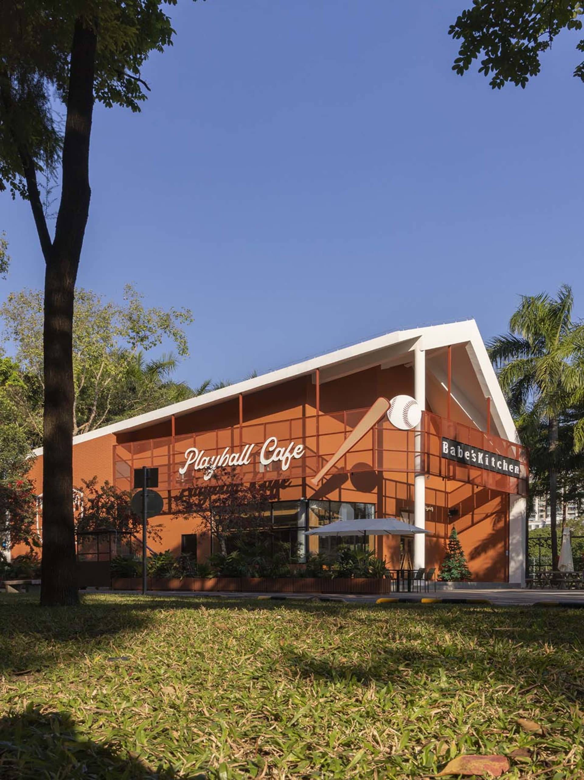 A modern restaurant with a large orange building, patio area, and potted plants, featuring a large window and well-lit interior