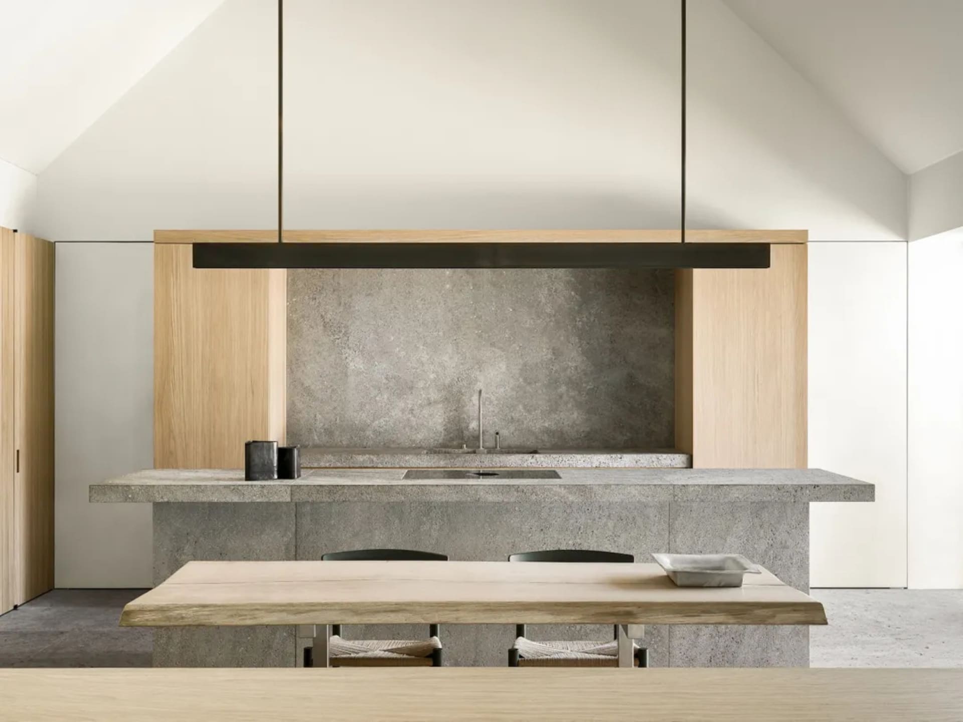 A modern kitchen with a wooden table and marble top, surrounded by black chairs, a sink, and a hanging light, with natural lighting creating a warm atmosphere