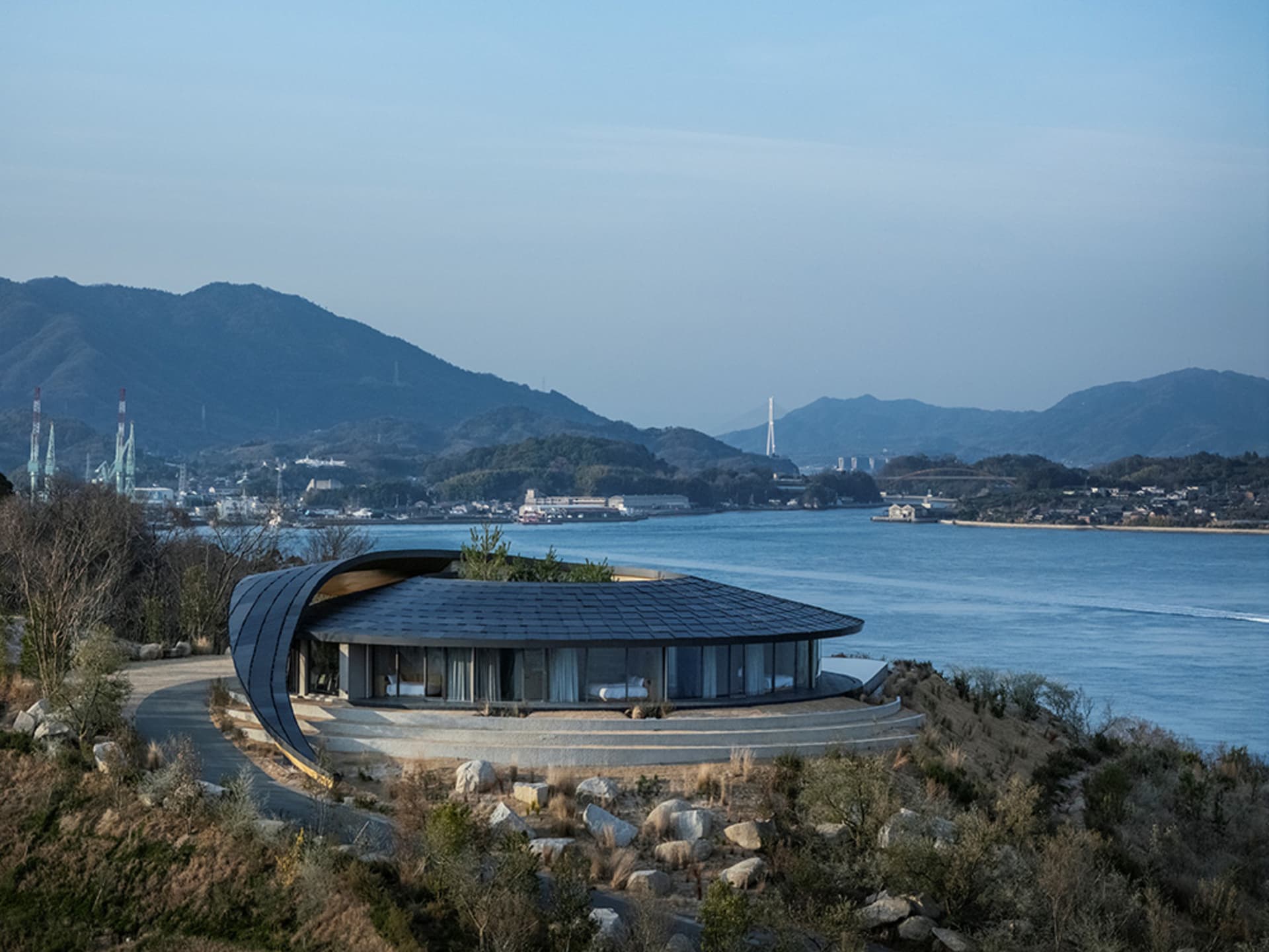 一座坐在山坡上遠眺水景的現代石材外觀住宅，屋頂為金屬屋頂，窗戶面積大，充滿自然光線