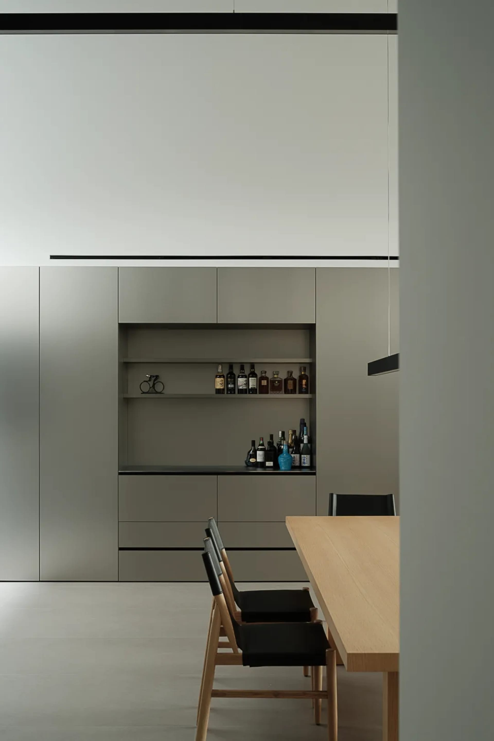 A modern kitchen with a wooden dining table and chairs, surrounded by bottles and a counter with a shelf, set against a dimly lit background