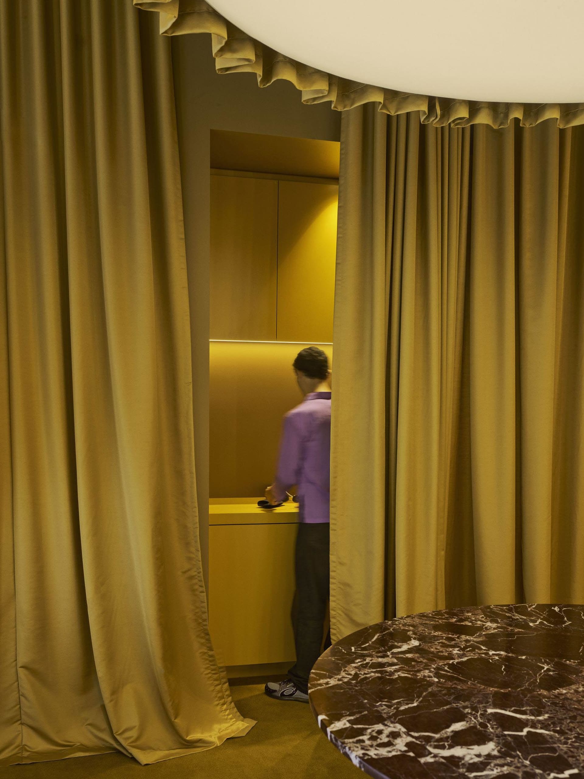 A dimly lit bathroom with a man using a cell phone, a yellow curtain, a sink, and a mirror