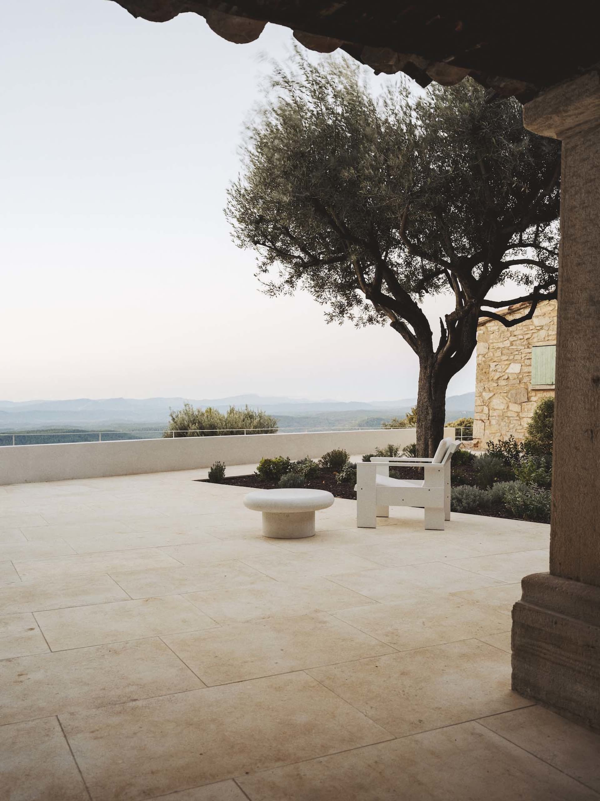 A relaxing outdoor patio with a stone wall, a white bench, and a wooden chair, surrounded by a tree and natural surroundings