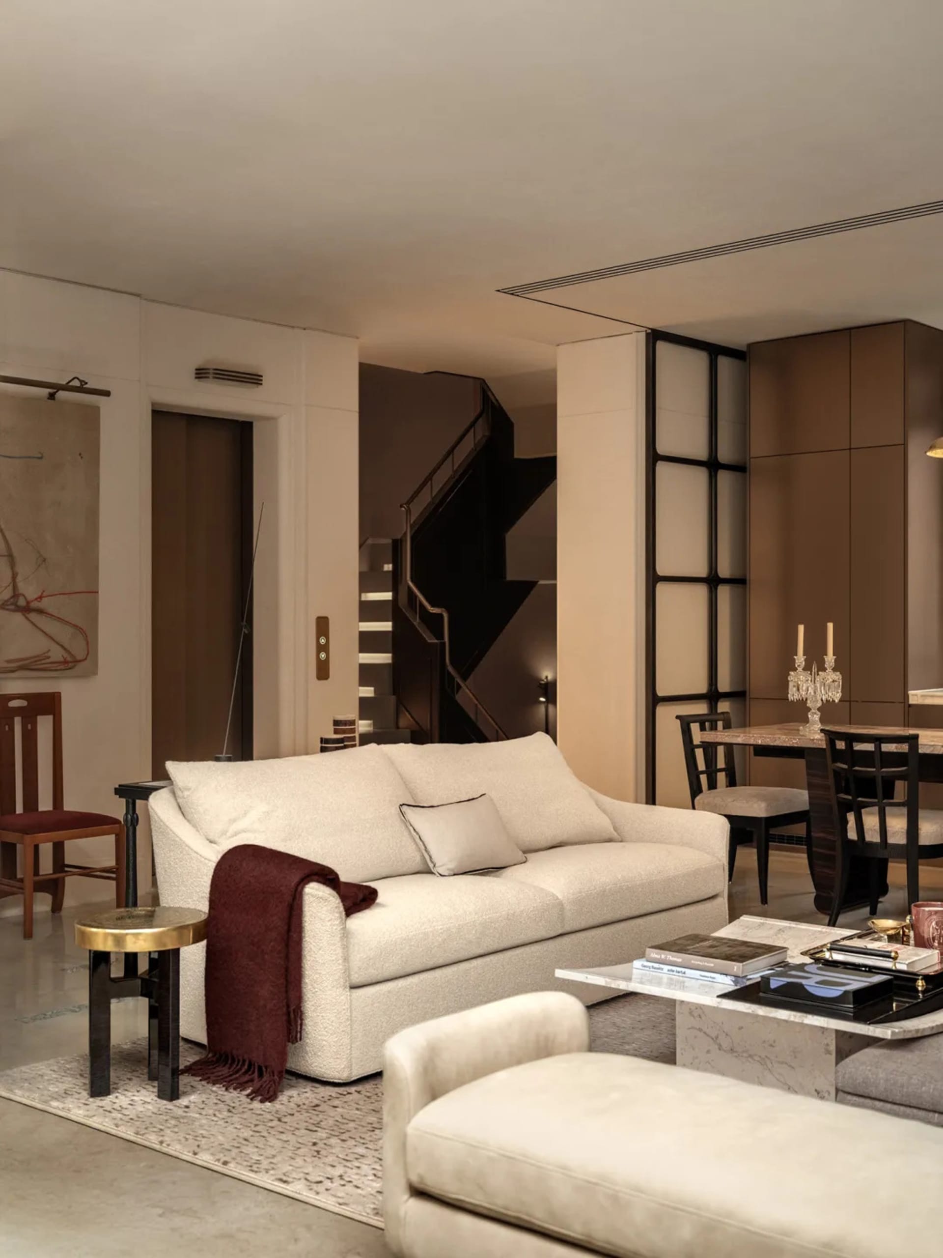 A well-lit living room with a white couch, a chair, and a dining table, featuring a staircase and a fireplace, with wood and white materials creating a clean and sophisticated atmosphere