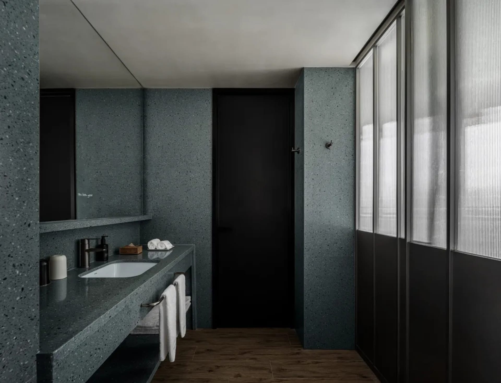 A modern hotel bathroom with a black door and countertop, featuring a sink under a mirror in a dimly lit space
