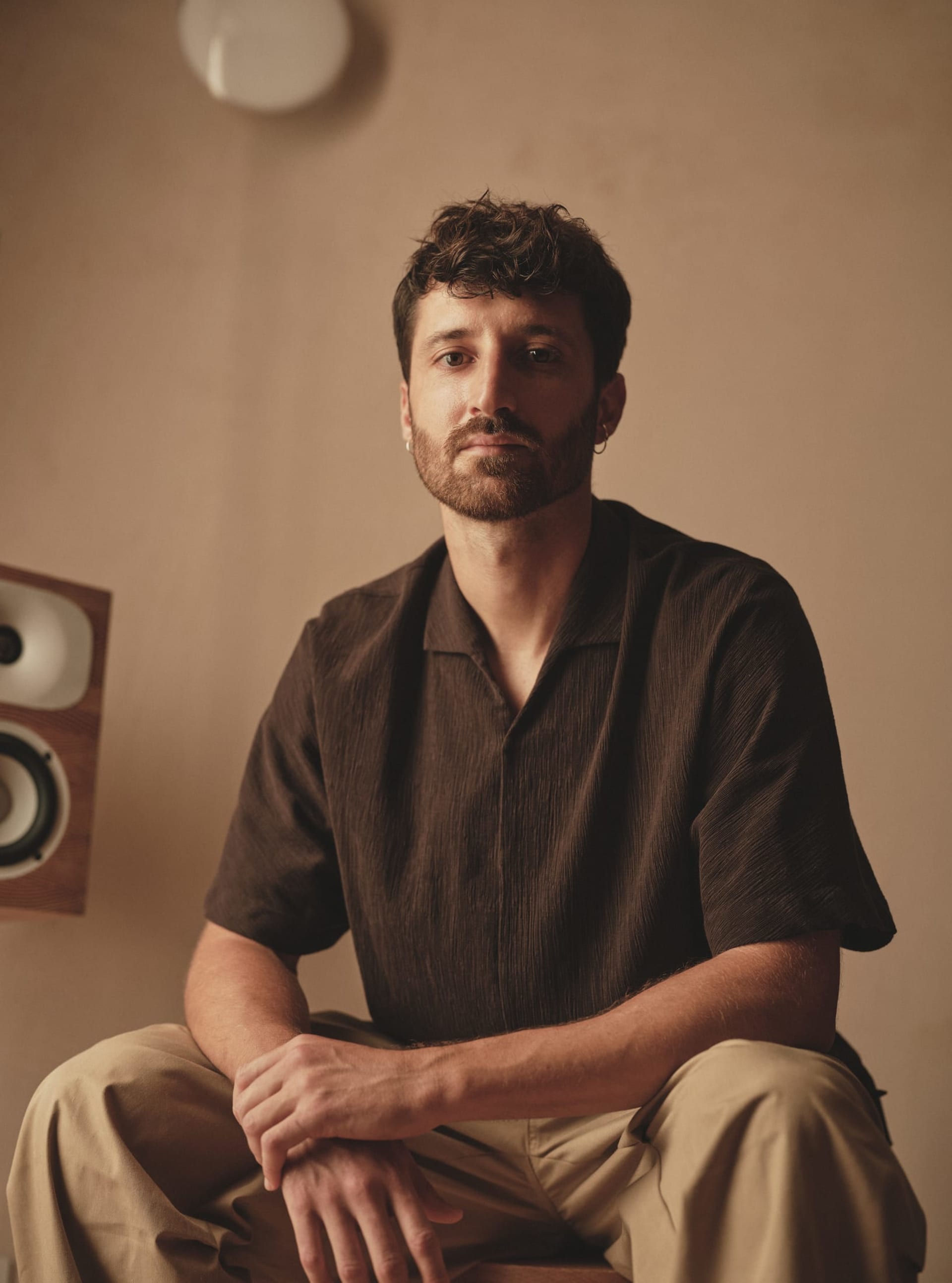 A man sitting on a wall, wearing a brown shirt, in a casual outdoor setting with speakers in the background