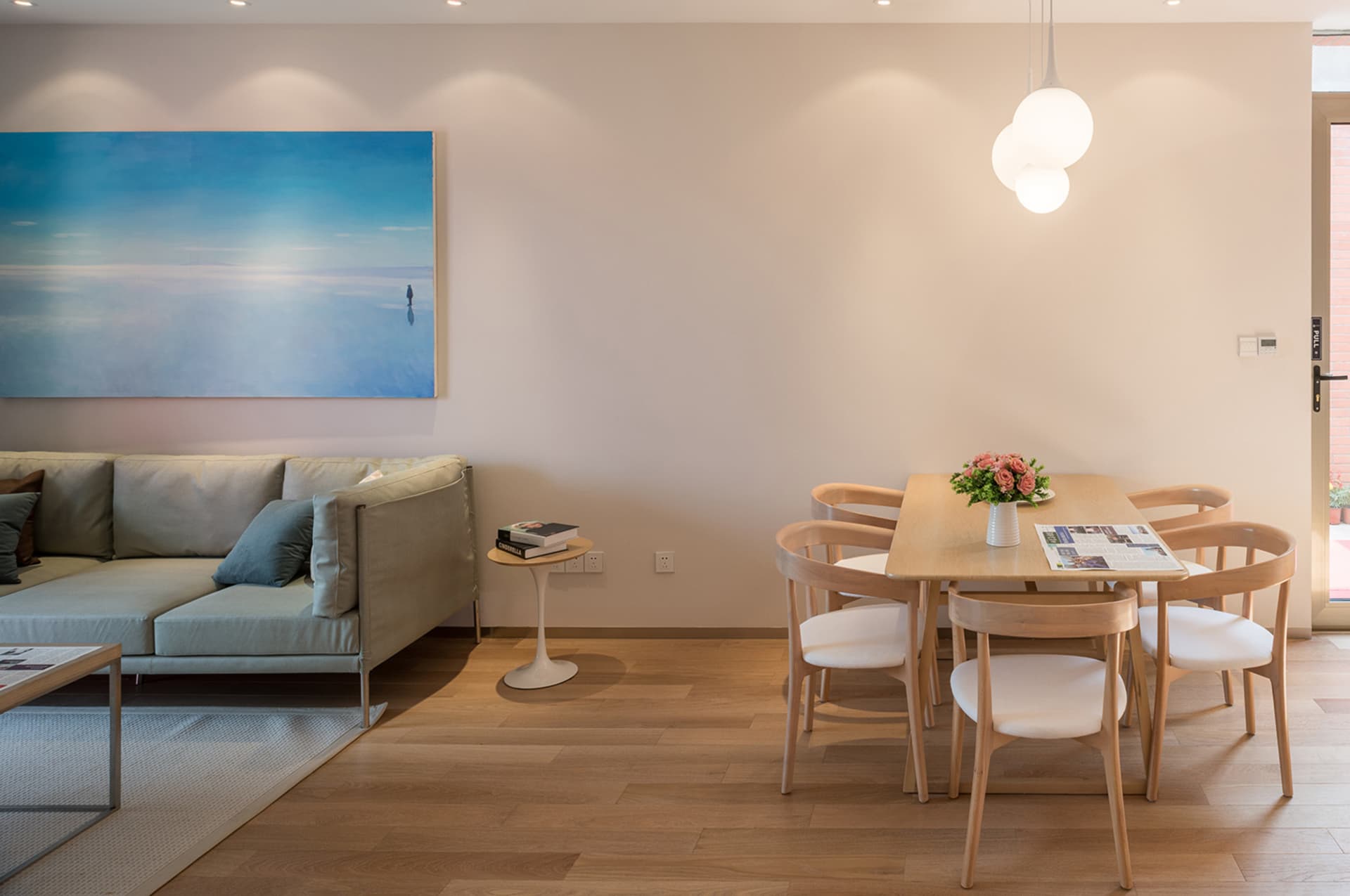 A warm and inviting living room with a wooden floor, a couch, a dining table, and a vase with flowers, surrounded by chairs and a potted plant