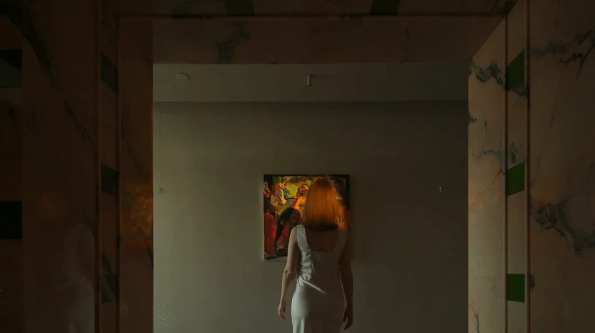A woman in a white dress admires a painting in a room with a white wall and brown floor