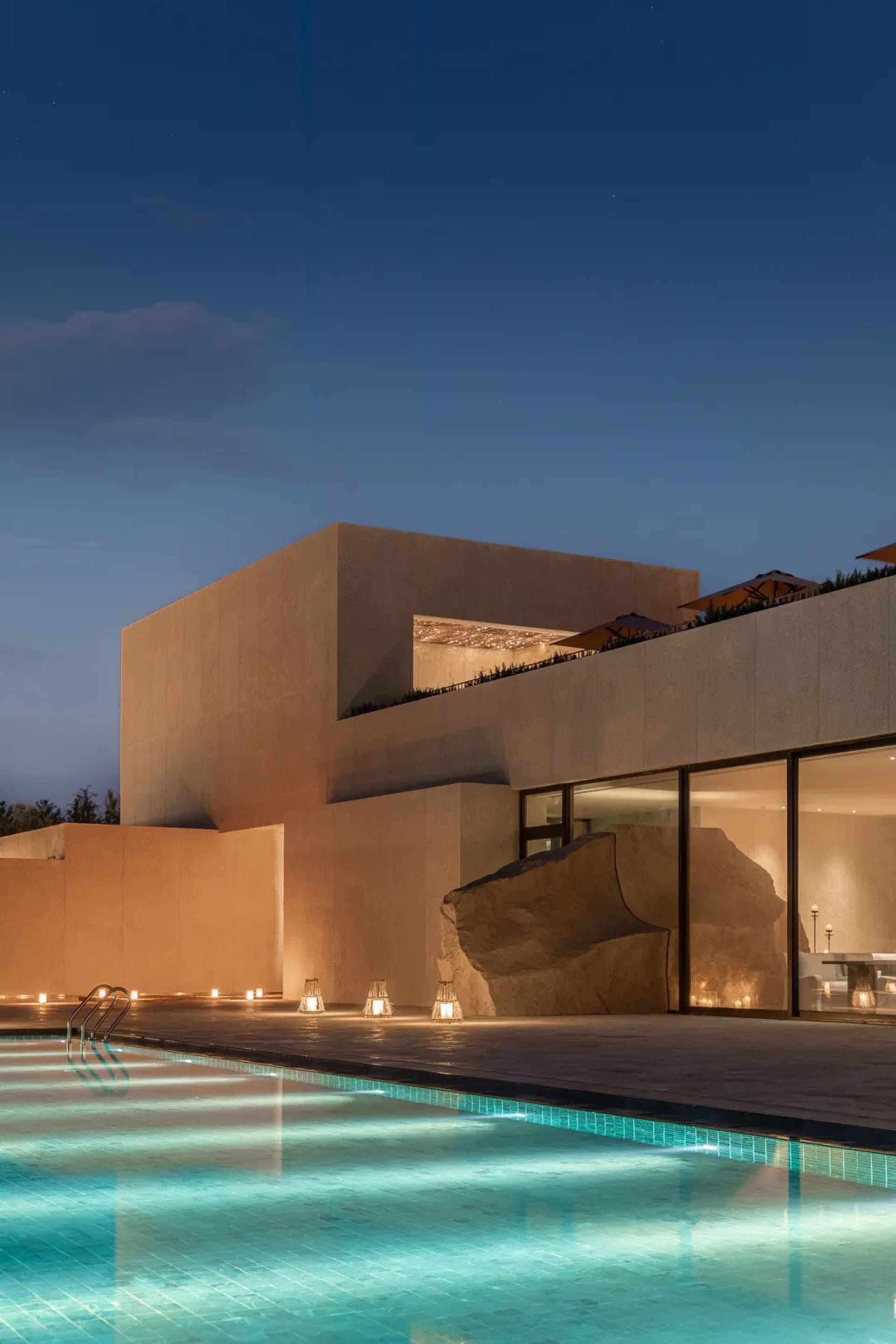 A modern house with a concrete exterior, a swimming pool, and a large rock in the living room, set in a dimly lit atmosphere