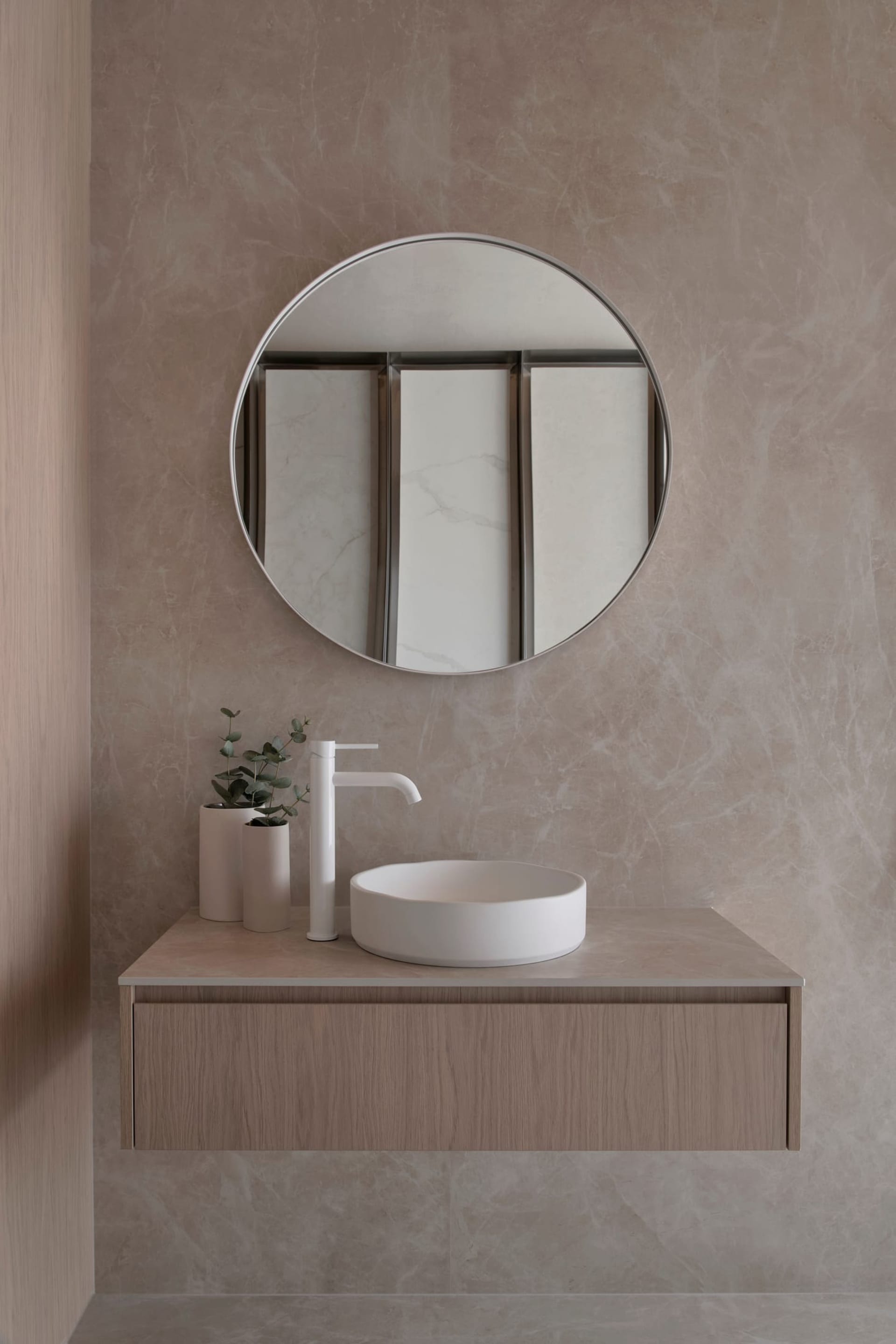 A modern bathroom with a white sink, large round mirror, and marble countertop, set against a wooden vanity, in a well-lit space