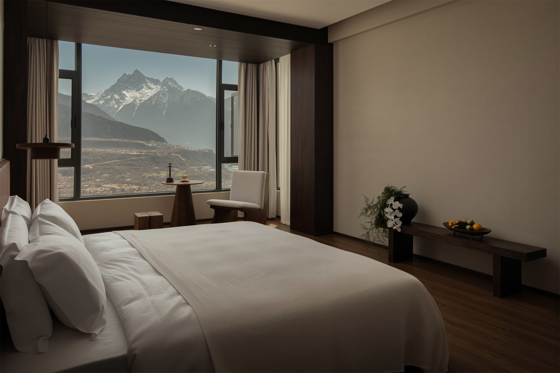 Cozy bedroom with a large window offering a view of mountains, featuring a white comforter, a chair, and a potted plant