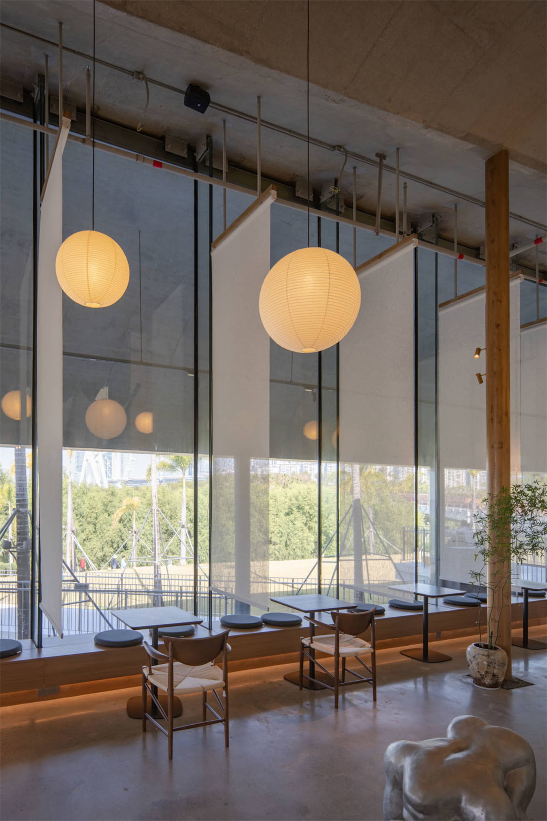A modern restaurant with a large window and glass wall, featuring comfortable seating, potted plants, and elegant decor, with a warm and welcoming atmosphere