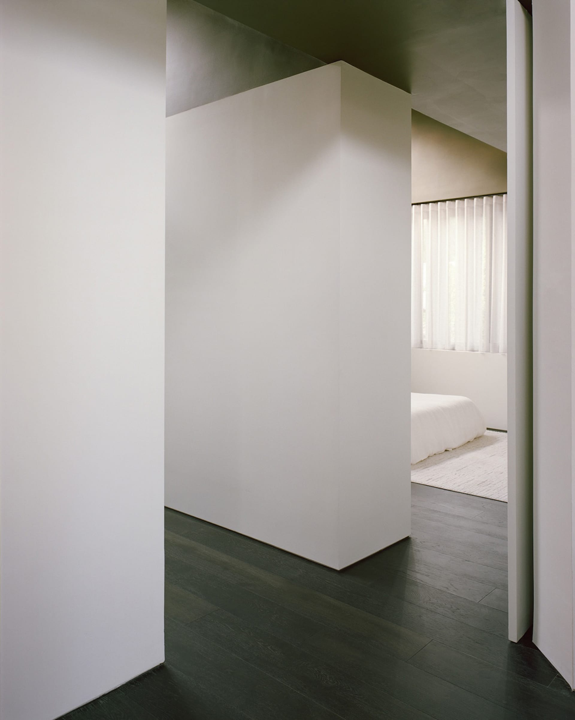 A well-lit bedroom with a white color scheme, featuring a bed near a window and a door on the right side
