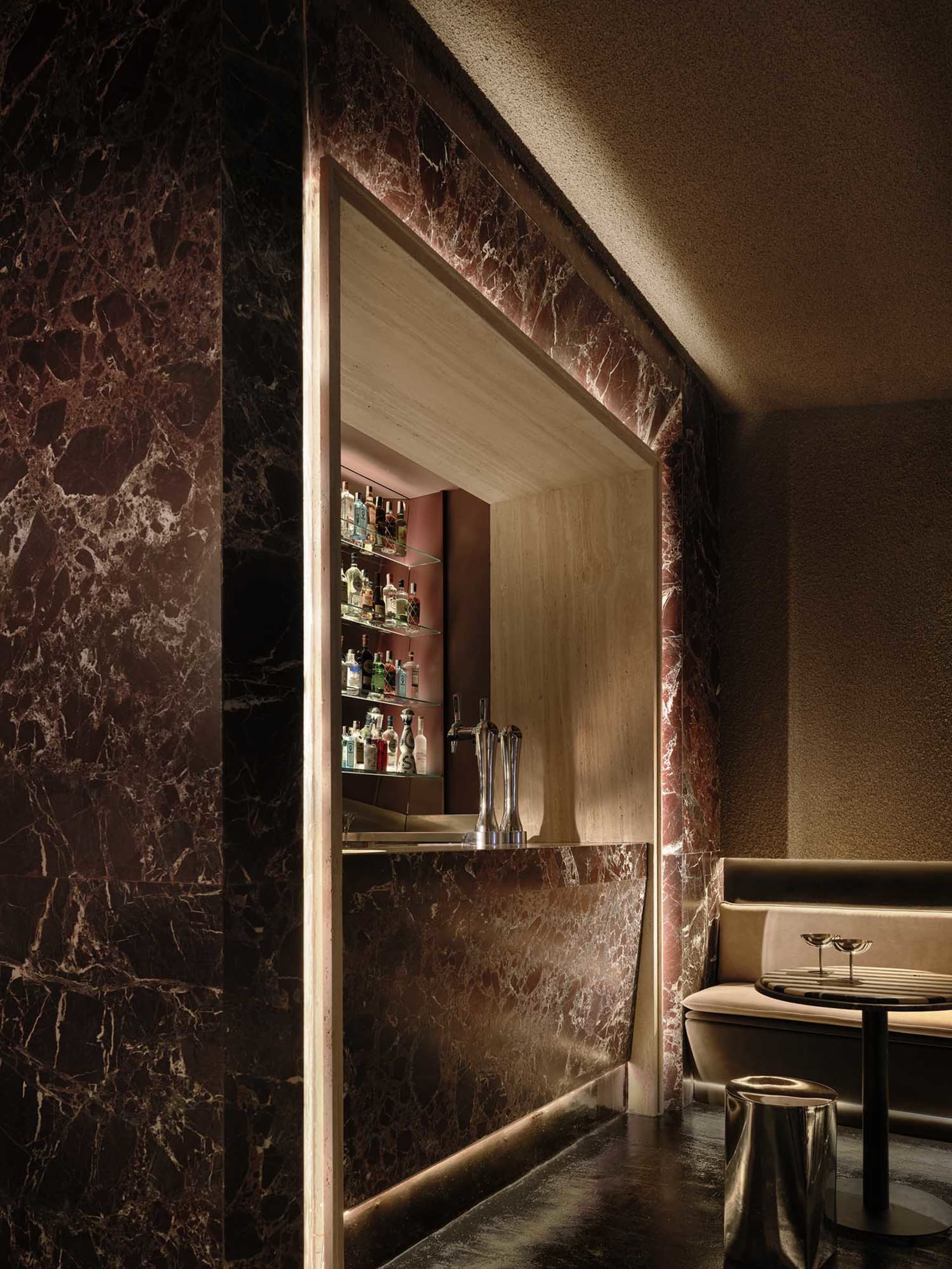 A dimly lit bar area with a marble bar, wooden counter, and bottles on display, set against a background of a sink and a couch in a modern and elegant restaurant