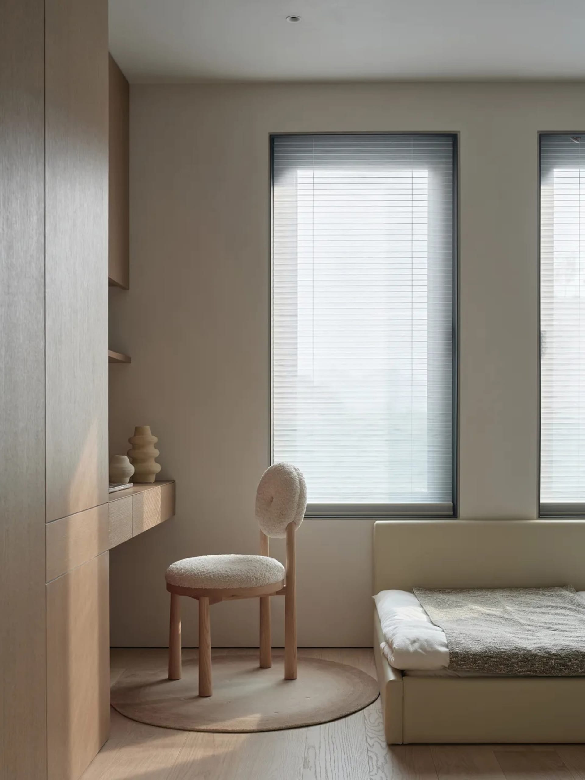 A bedroom with a bed, chair, and window, featuring a simple and clean design with white materials and natural light