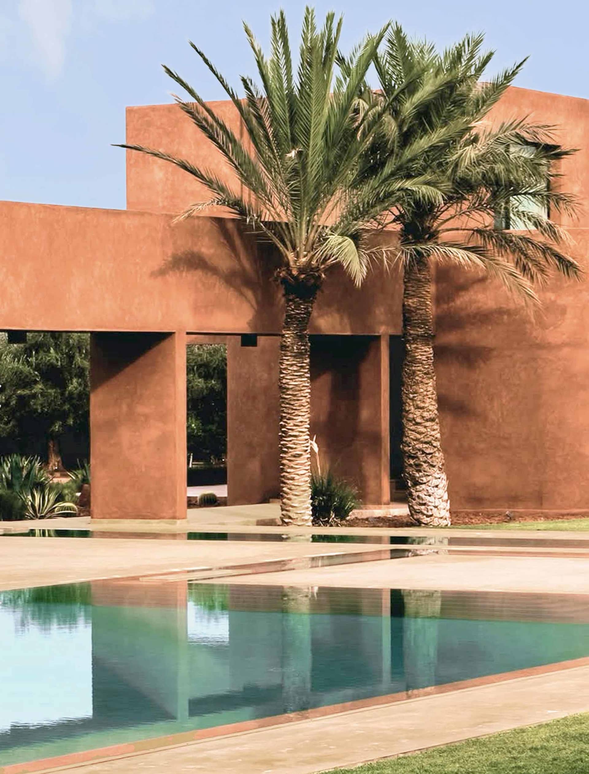 A serene desert pool area with a blue pool, palm trees, and a red building in the background