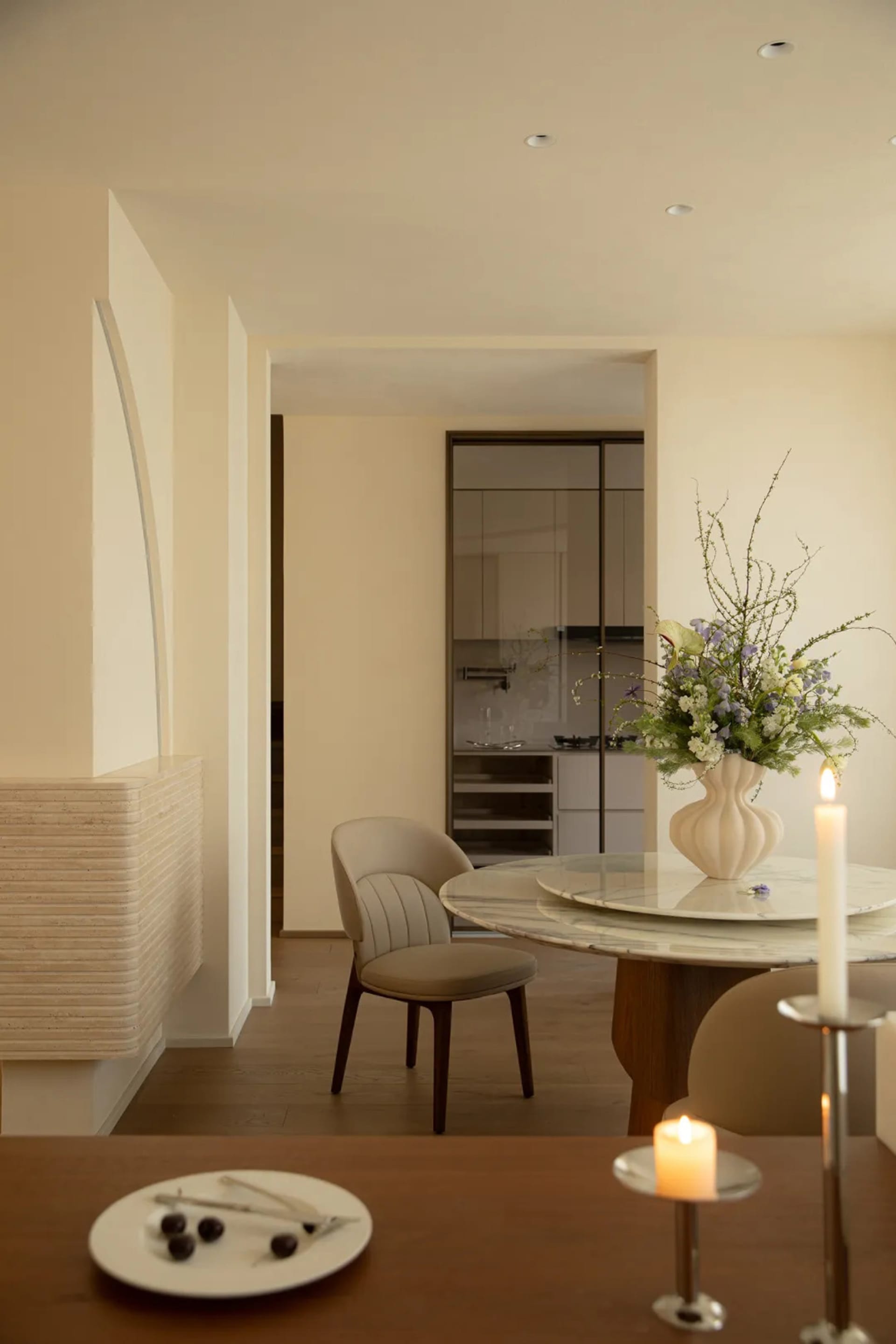 Cozy living room with a wooden floor, a dining table, a couch, and a chair, with a dimly lit atmosphere and a vase with flowers on the table