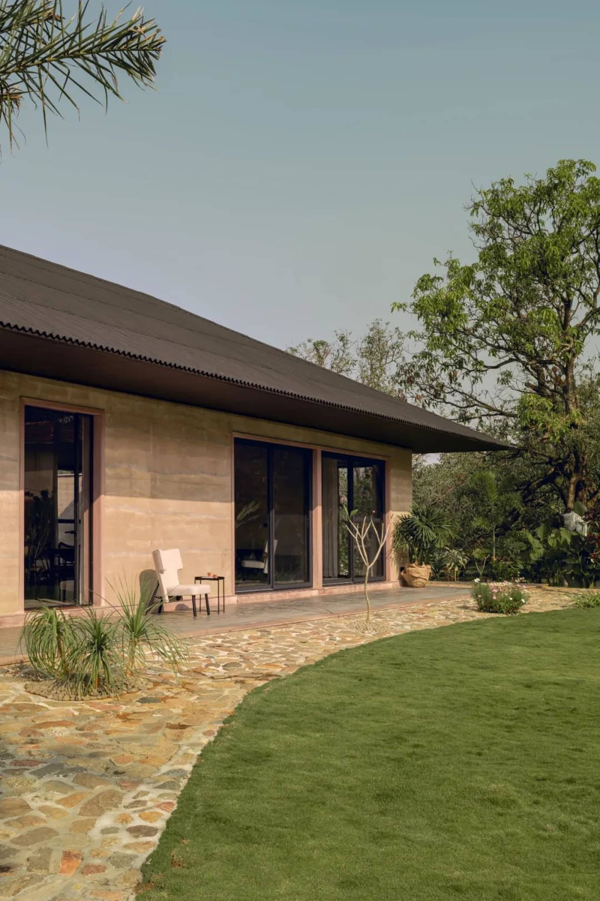 Photograph of a house with a stone exterior, a black roof, a patio with a dining table and chairs, and a garden with a tree and a bench
