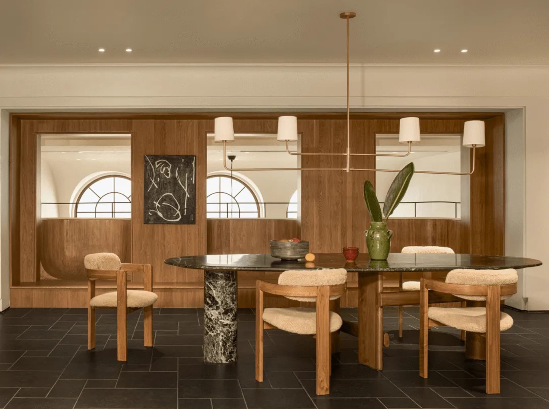 A modern dining room with a large wooden table and chairs, decorated with a bowl, vase, and potted plant, surrounded by natural light from the windows
