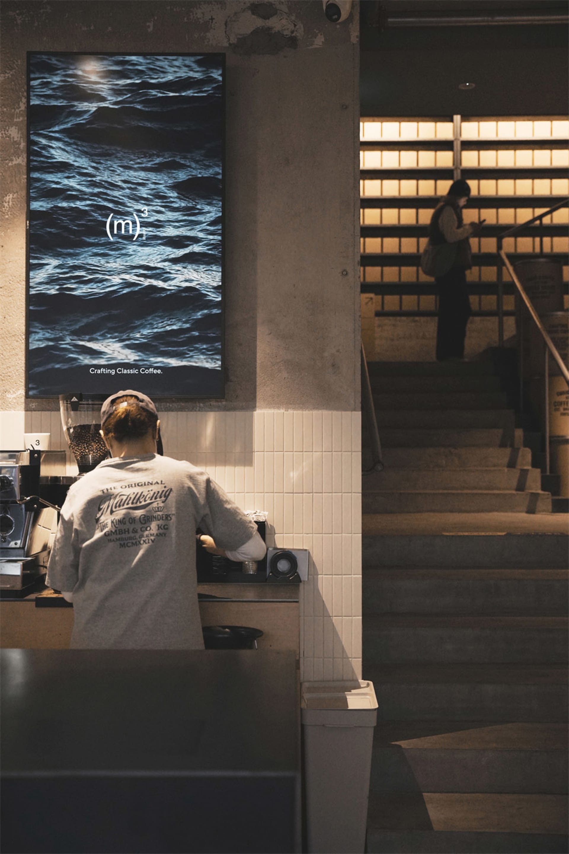 A woman stands behind a wooden counter in a cozy coffee shop with a staircase and books on the shelves