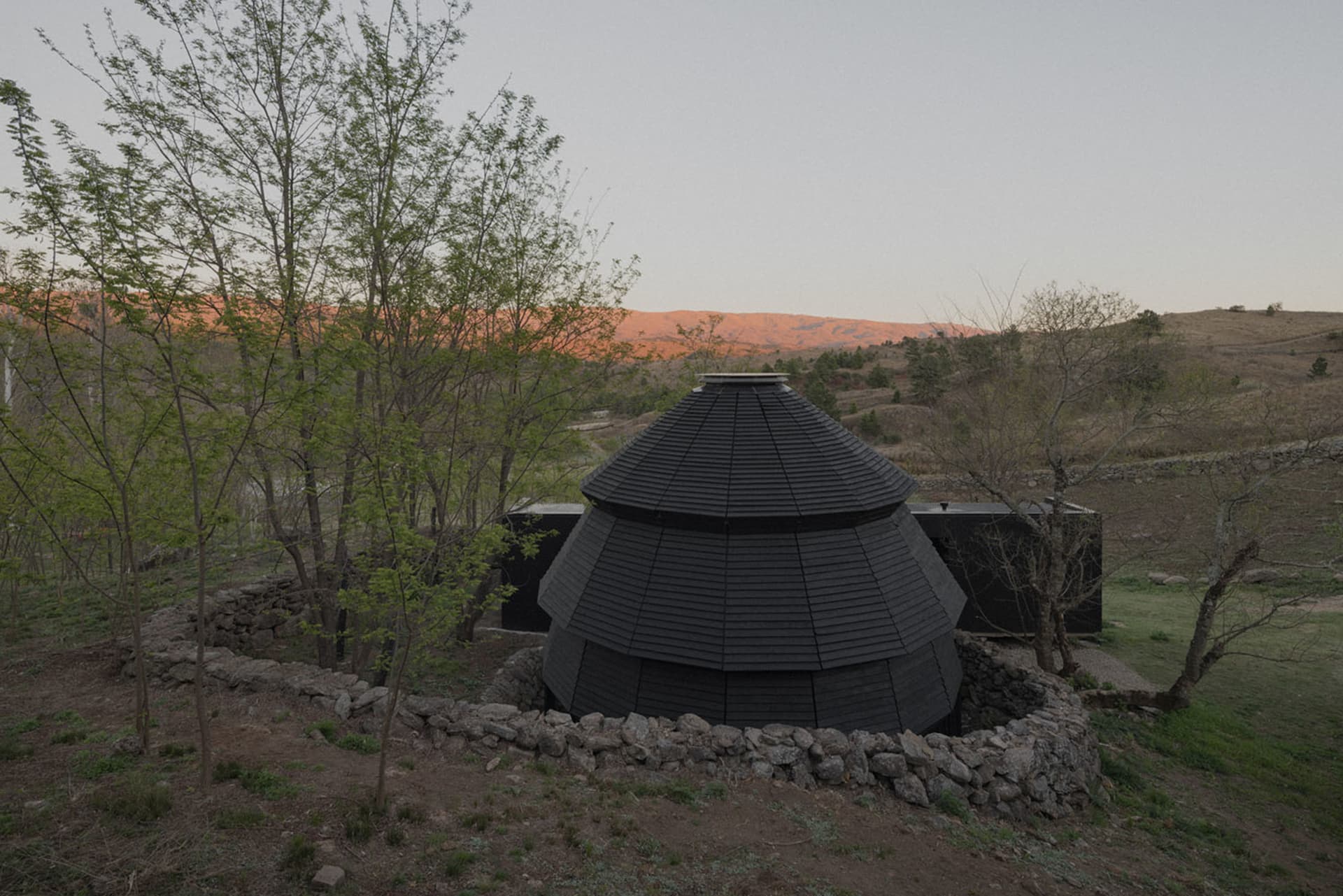 A rustic house with a black roof and a circular structure on top, situated in a wooded area during sunset