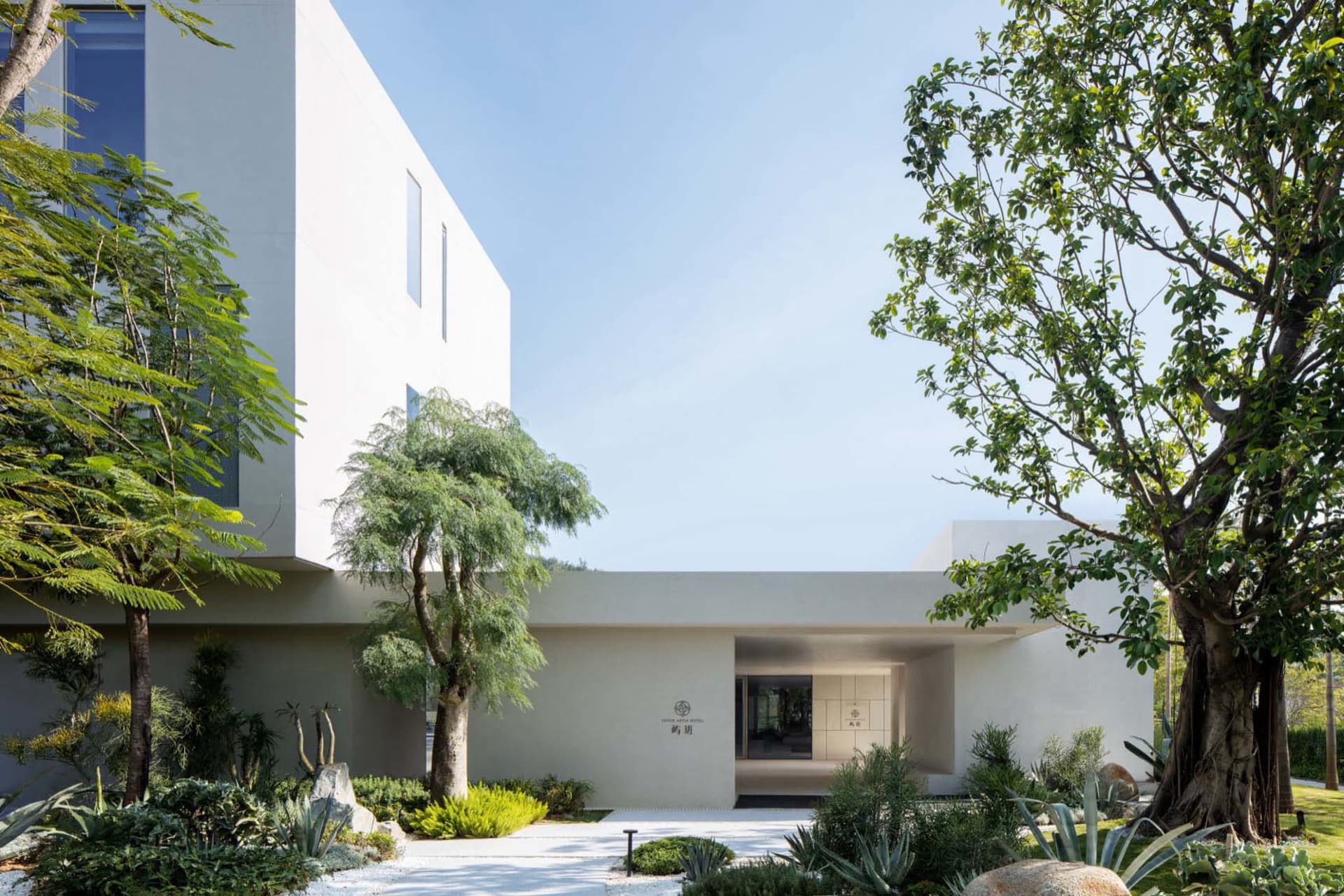 A modern house with a white exterior, large window, and balcony, surrounded by trees and a potted plant, with natural light streaming in through the window