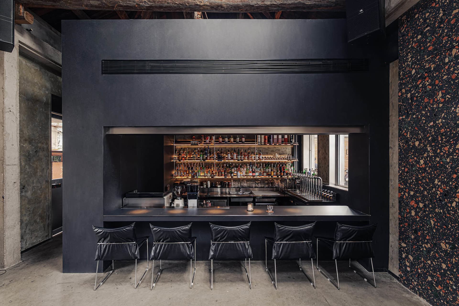A dark and intimate bar with a long wooden counter and numerous bottles, surrounded by chairs for patrons