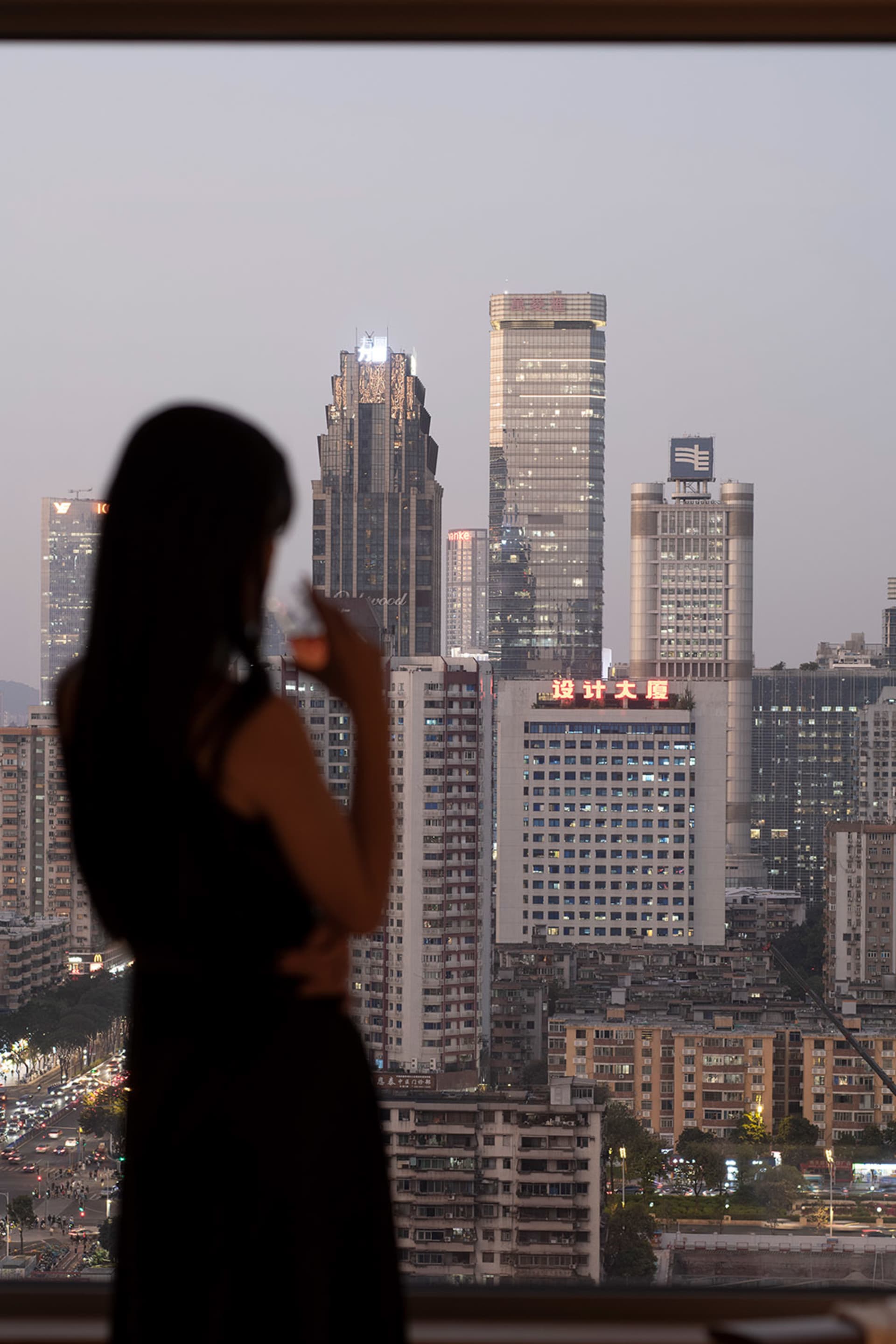 A woman stands on a rooftop at night, admiring the city's vibrant and lively atmosphere, illuminated by artificial lights