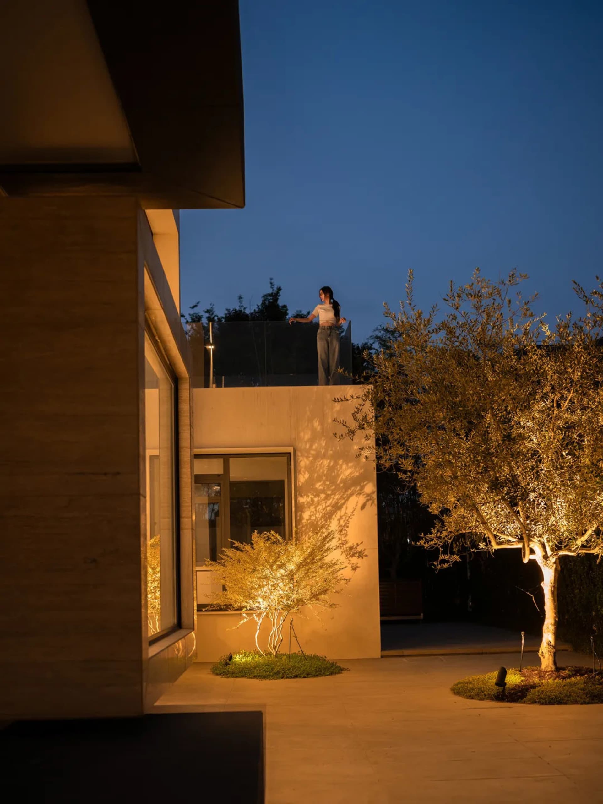 A woman stands on a modern house balcony at night, surrounded by city lights and streetlights, creating a warm and inviting atmosphere