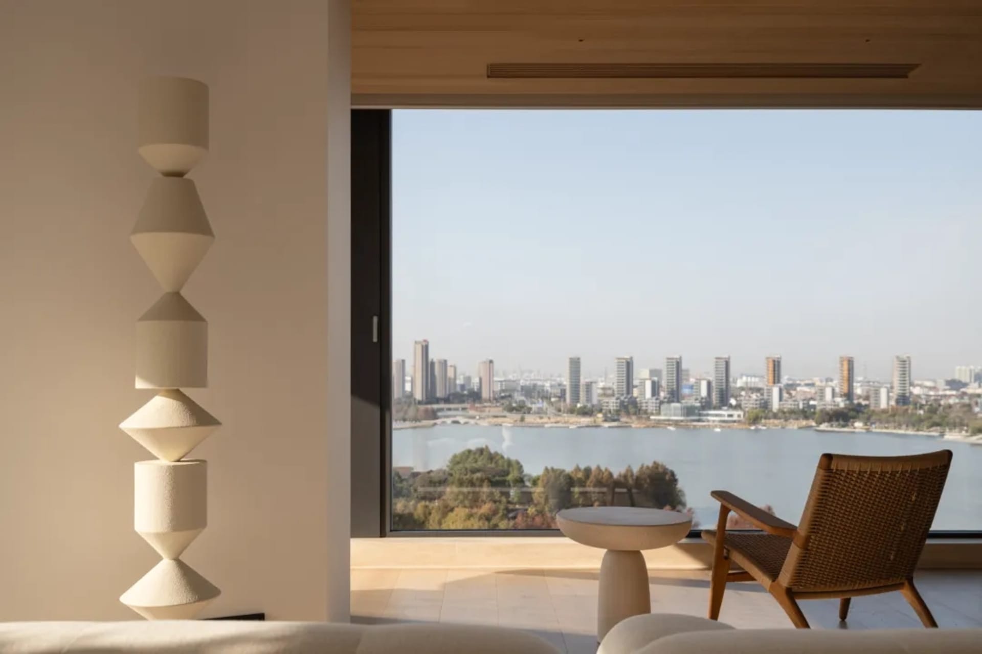 A modern living room with a couch, chair, and dining table, featuring a large window with a city view, wood and glass elements, and natural lighting