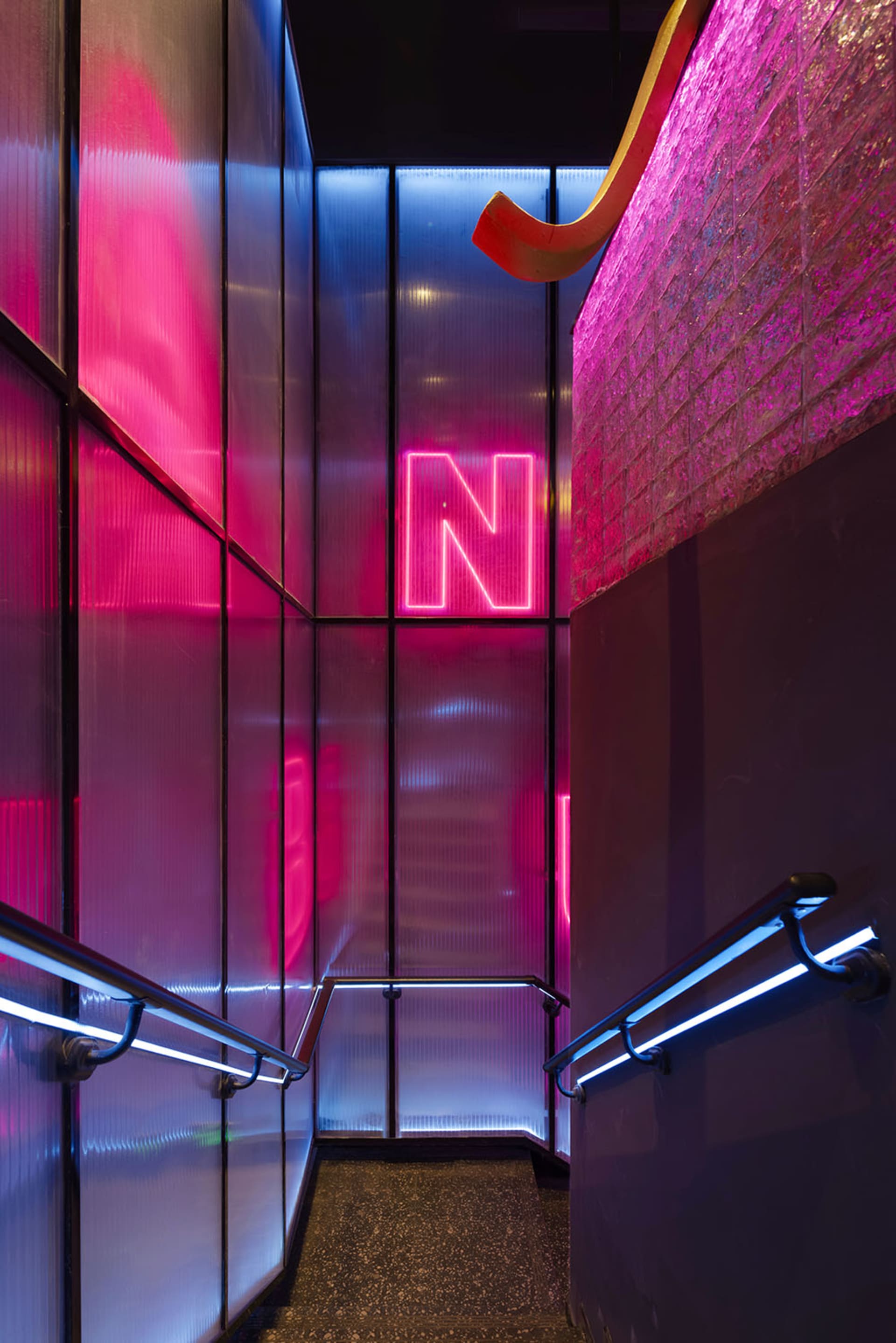 A dimly lit stairwell with a pink and red color scheme, featuring metal stairs and a red and white sign on the wall