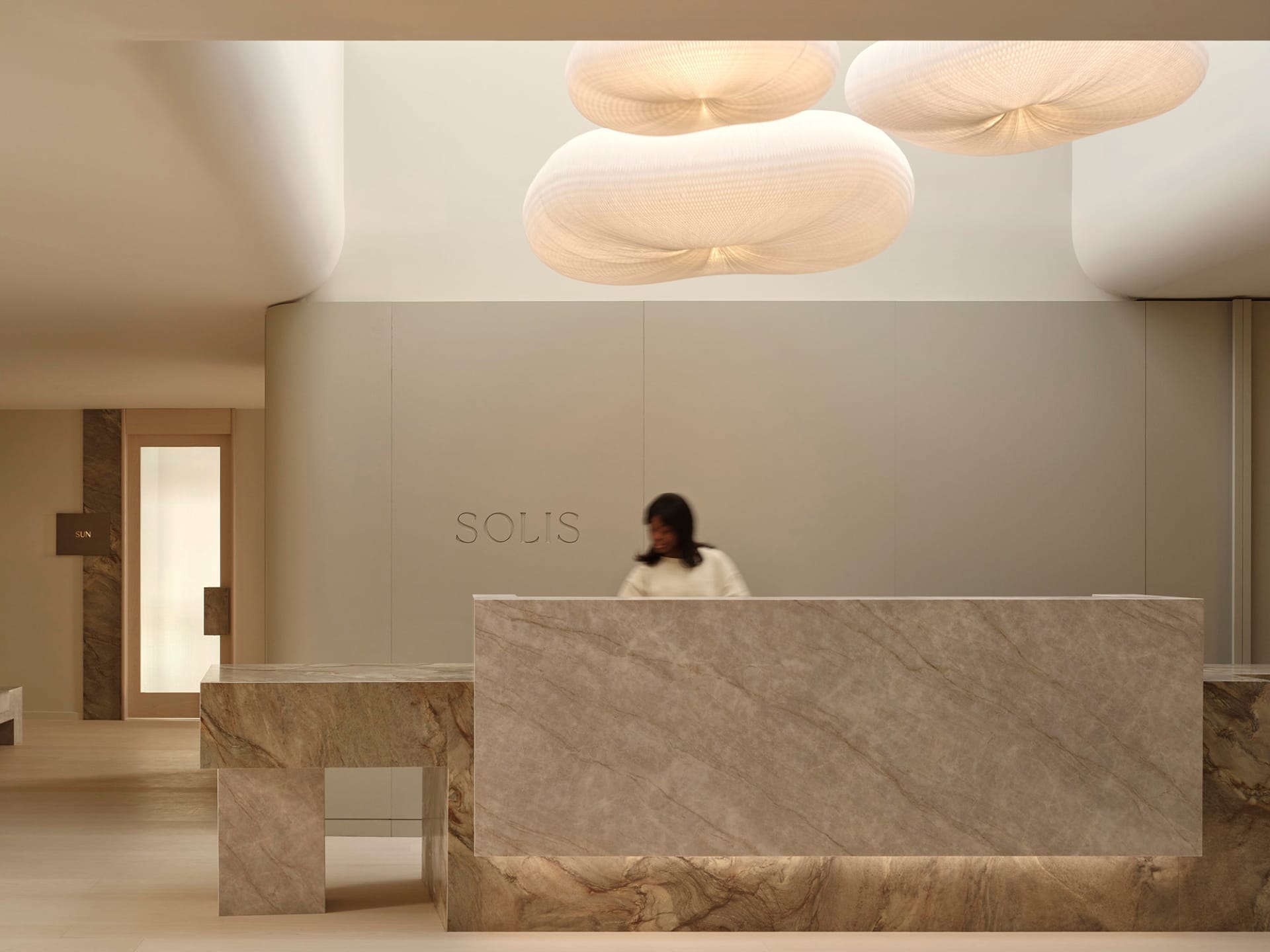 A modern lobby with a white marble countertop, grey marble wall, and a woman standing in front of the counter, with a nearby bench in a soft and inviting atmosphere
