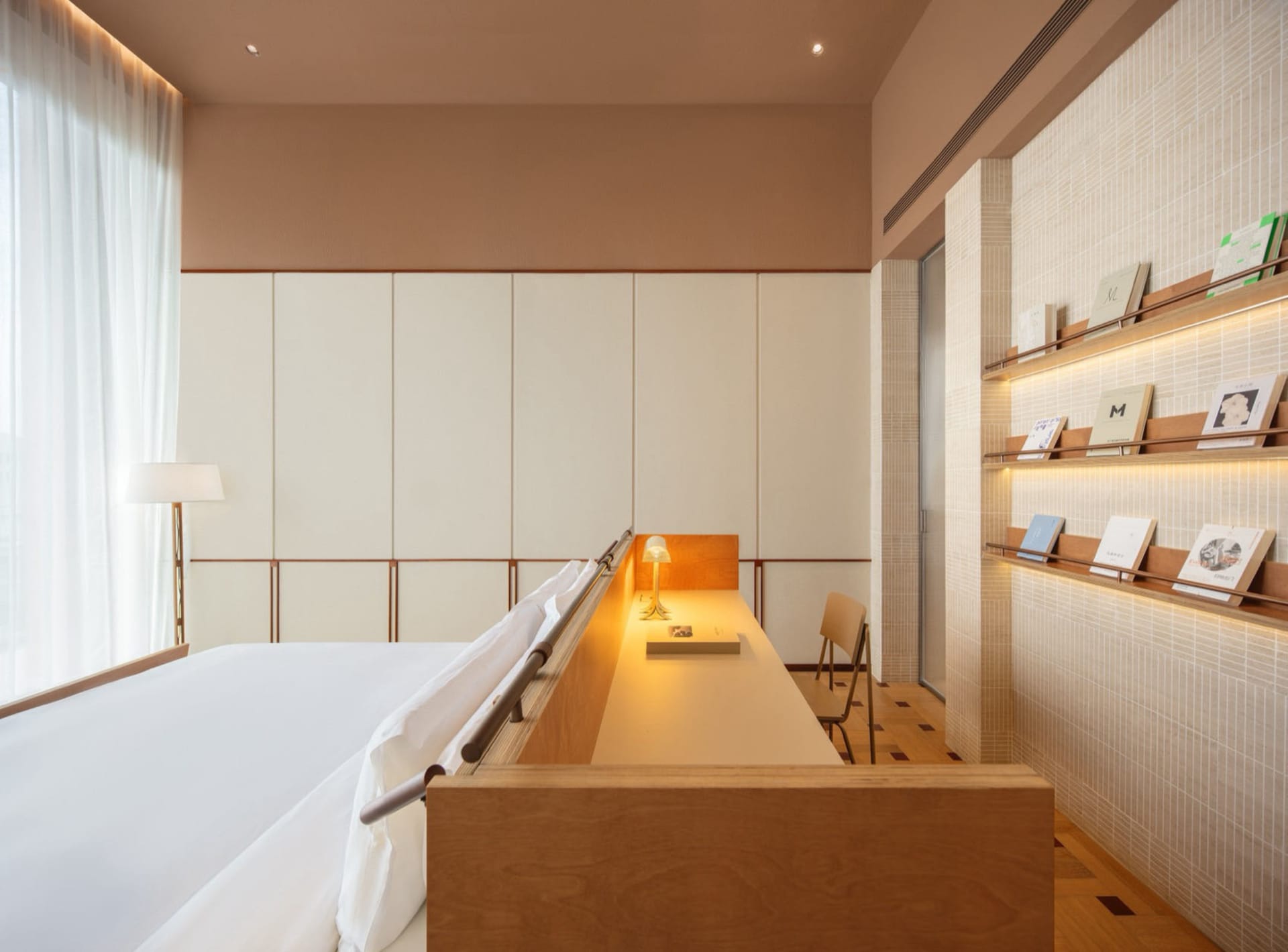 A modern bedroom with a bed and desk, featuring wood and white sheets, in a well-lit space with natural light from a window