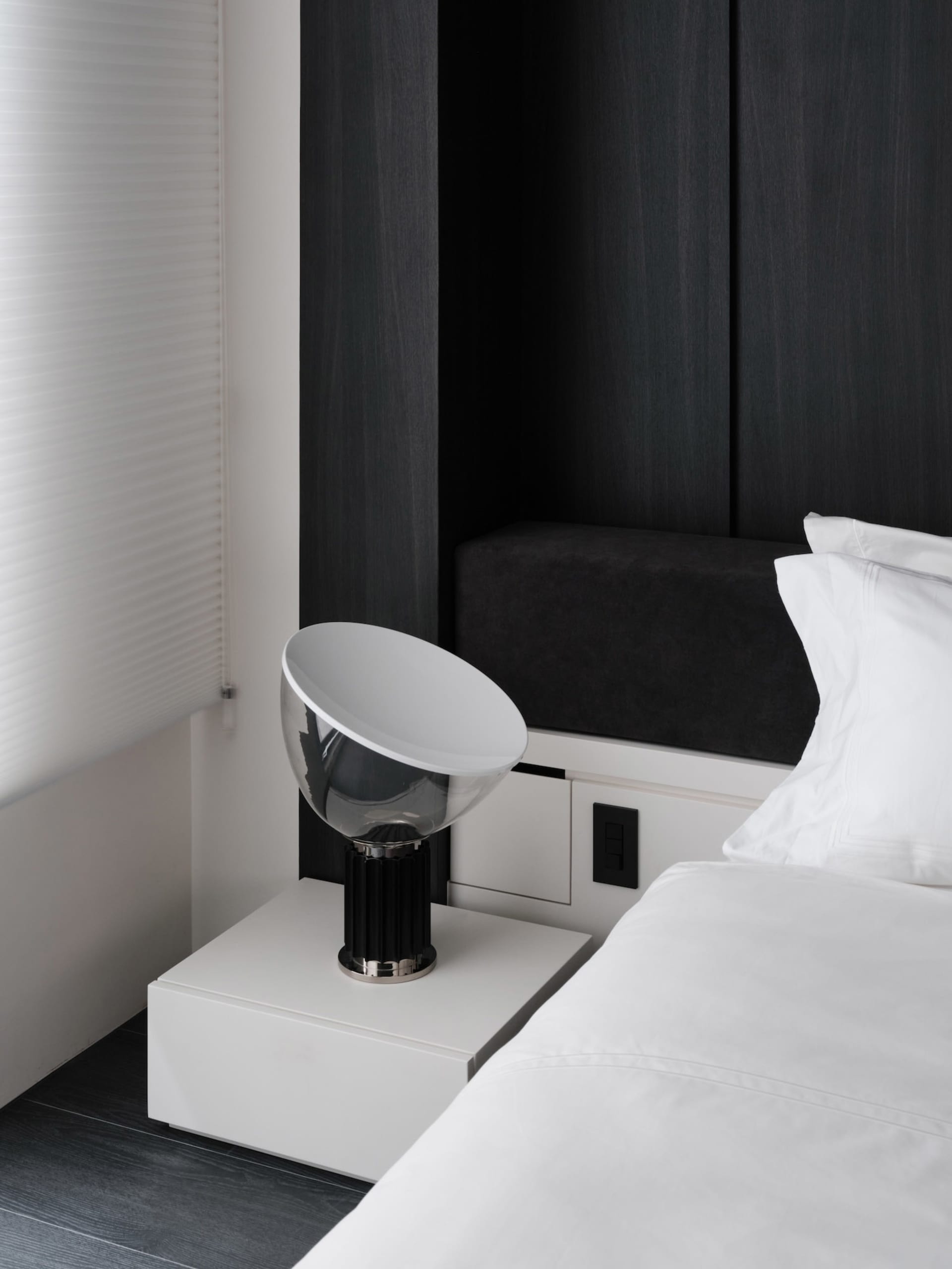 A dimly lit bedroom with a bed on the right, a bench on the left, and a table with a lamp in the middle