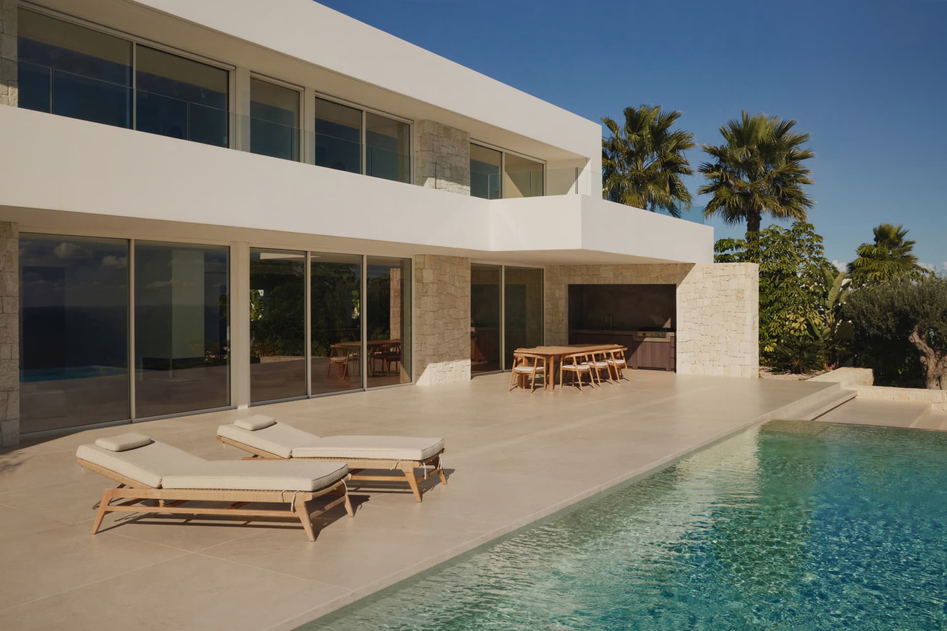 A modern house with a white exterior, a large patio, and a swimming pool surrounded by a deck and palm trees, with natural light and a relaxed atmosphere