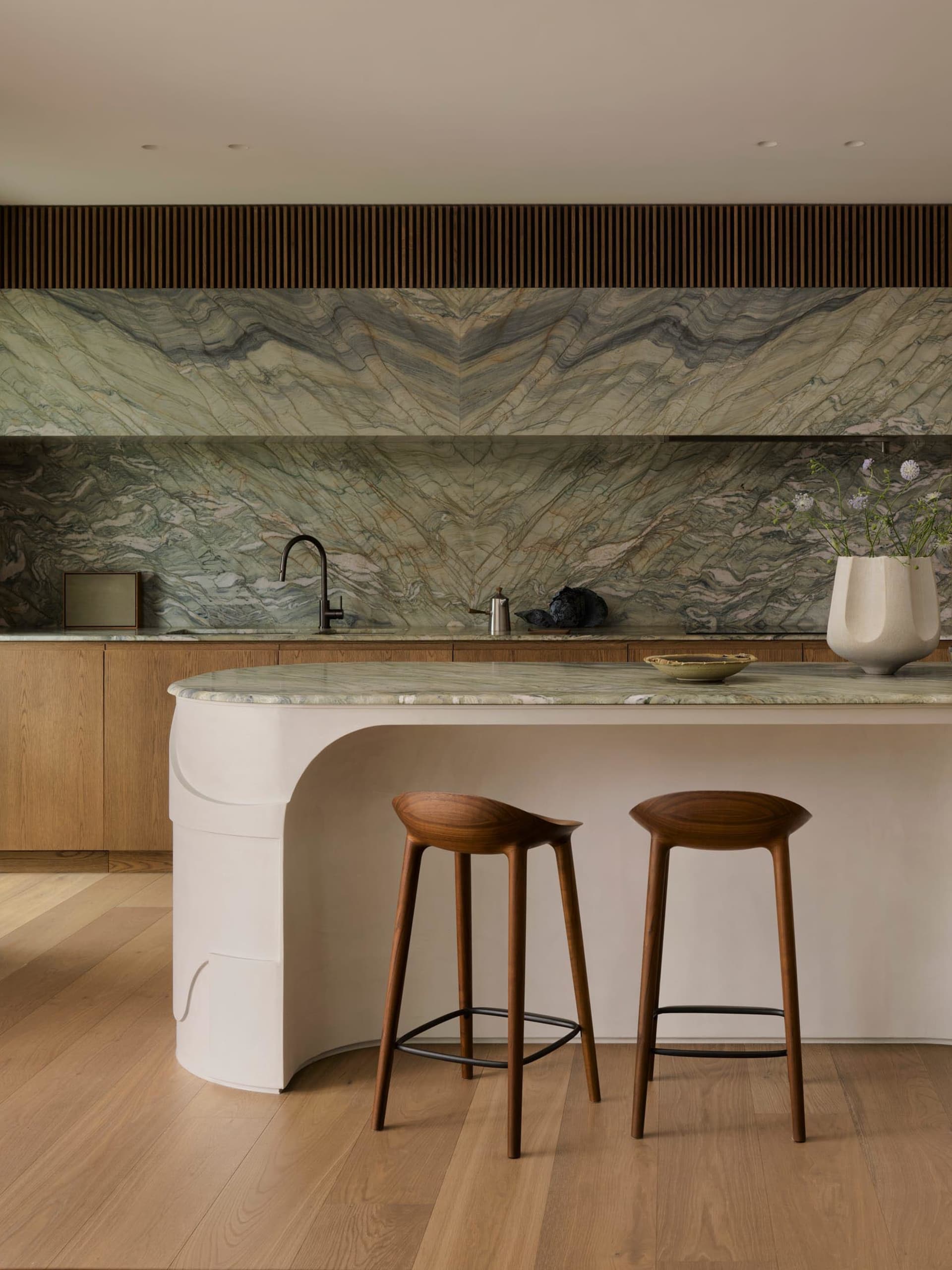 A dimly lit kitchen with a marble countertop, sink, and backsplash, accompanied by wooden stools and a potted plant, creating a cozy atmosphere