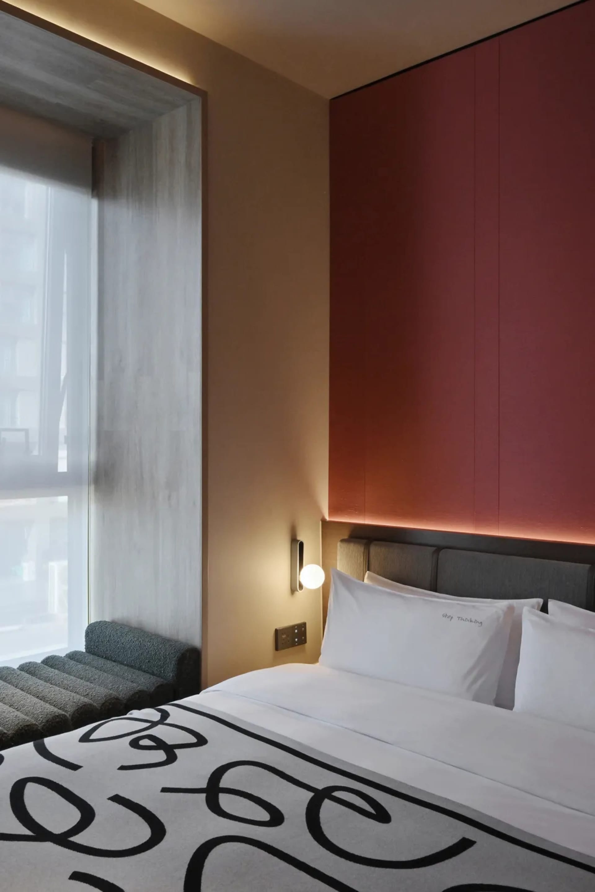 A modern hotel bedroom with a white bed, a bench, and a window, featuring a predominantly white color scheme and natural lighting