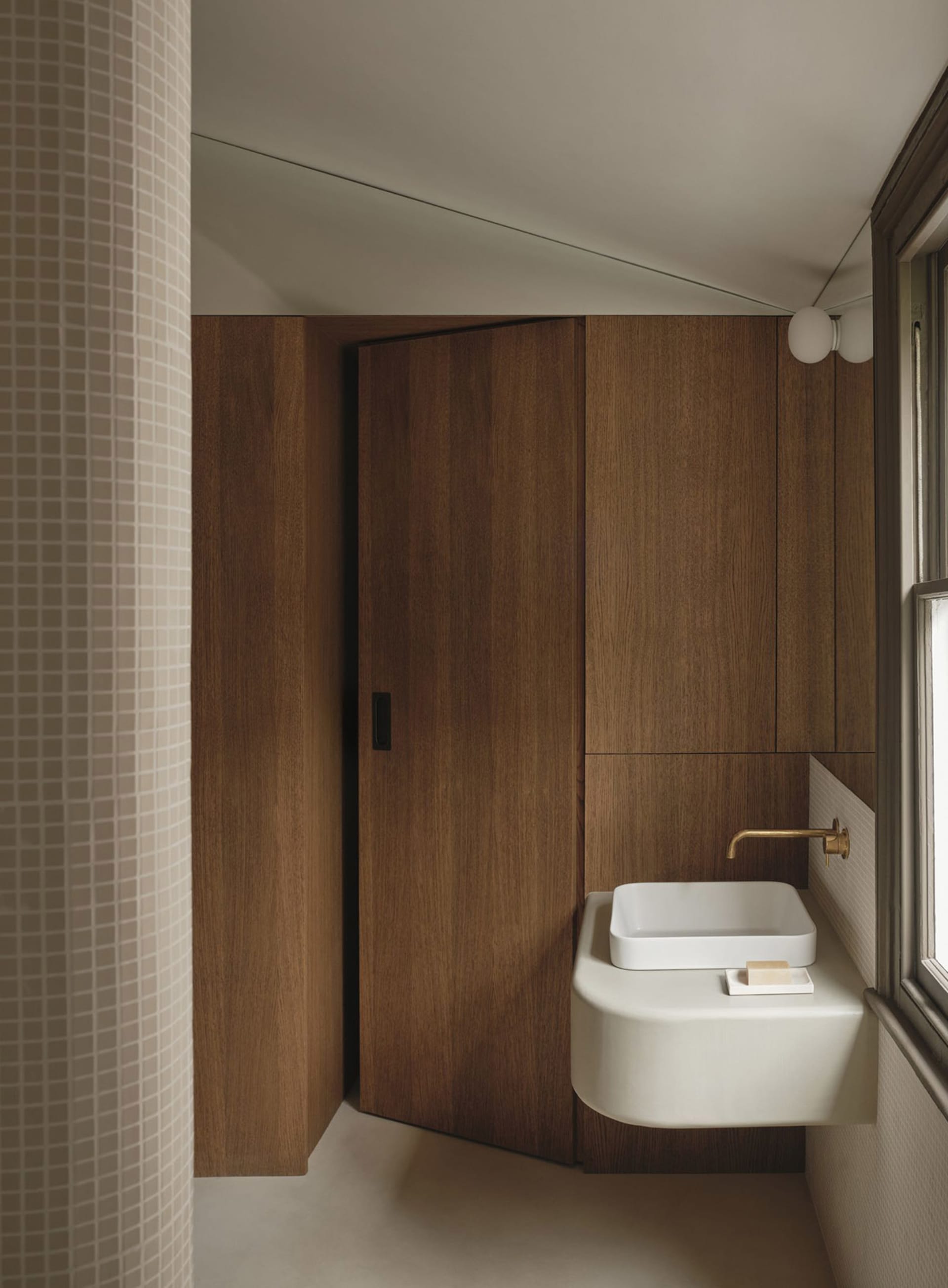 A modern bathroom with a white sink under a window and a brown wooden door, well-lit by natural light