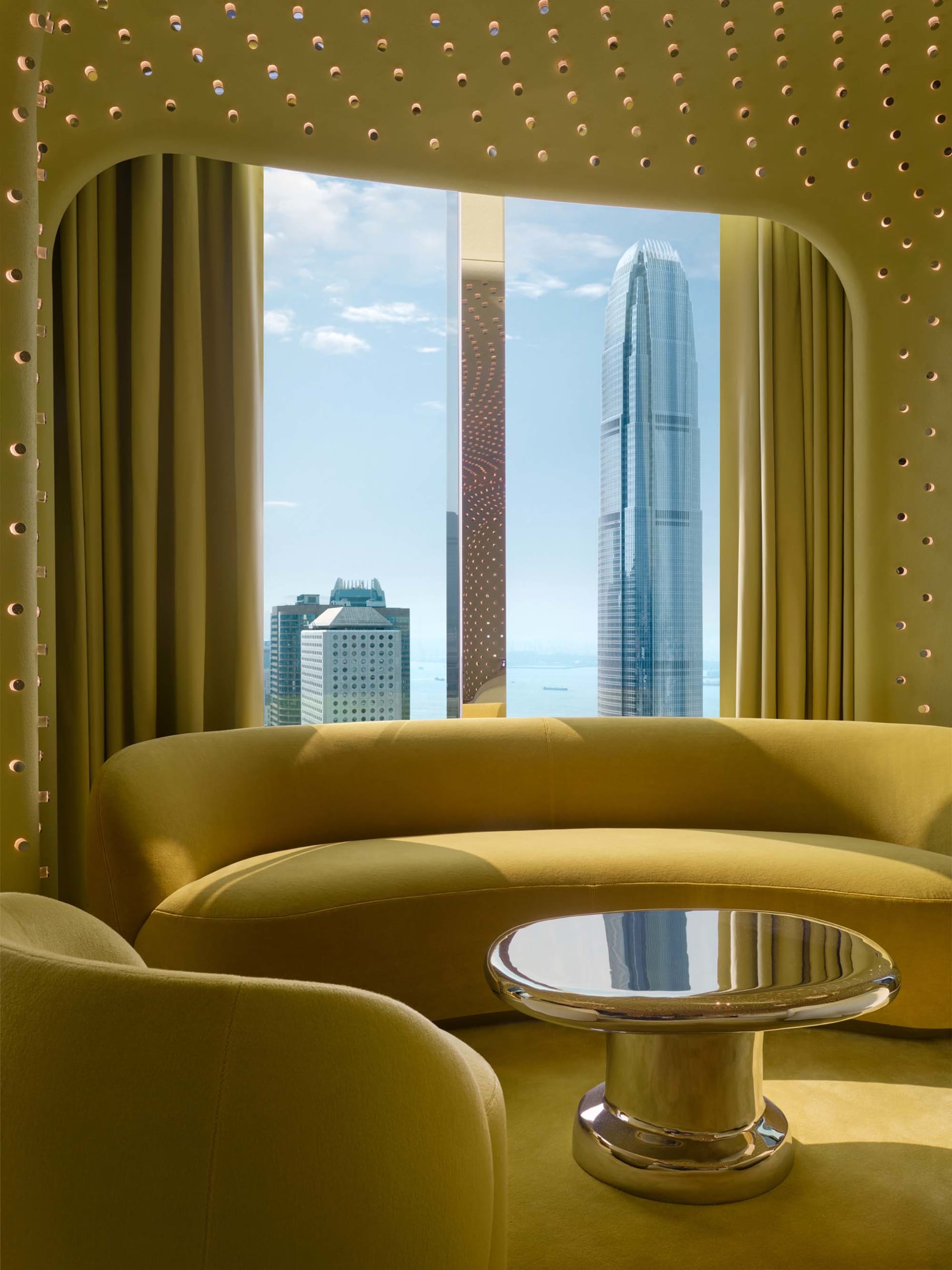 A well-lit living room with a couch and dining table, featuring a large window with a city view, wood furniture, and glass accents