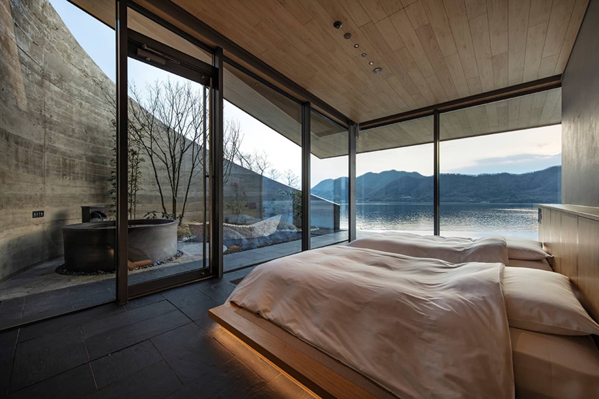 A modern hotel bedroom with a large bed, wooden headboard, and natural light from a large window