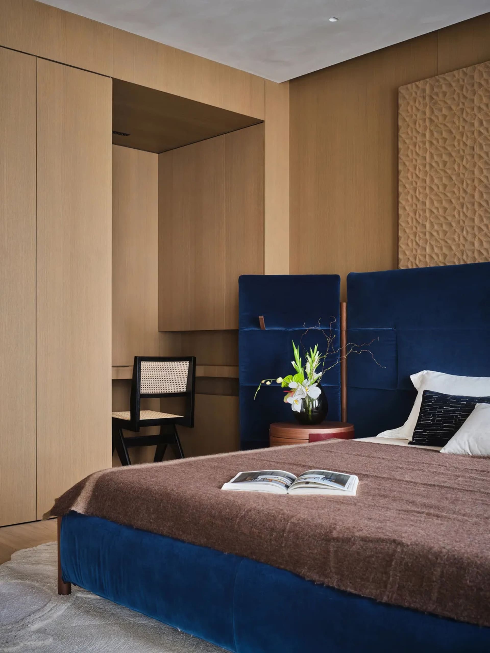 Cozy bedroom with a blue-covered bed, a wooden floor, and a brown color scheme, with a vase and flowers on a table, and a book on the bed, in a dimly lit space