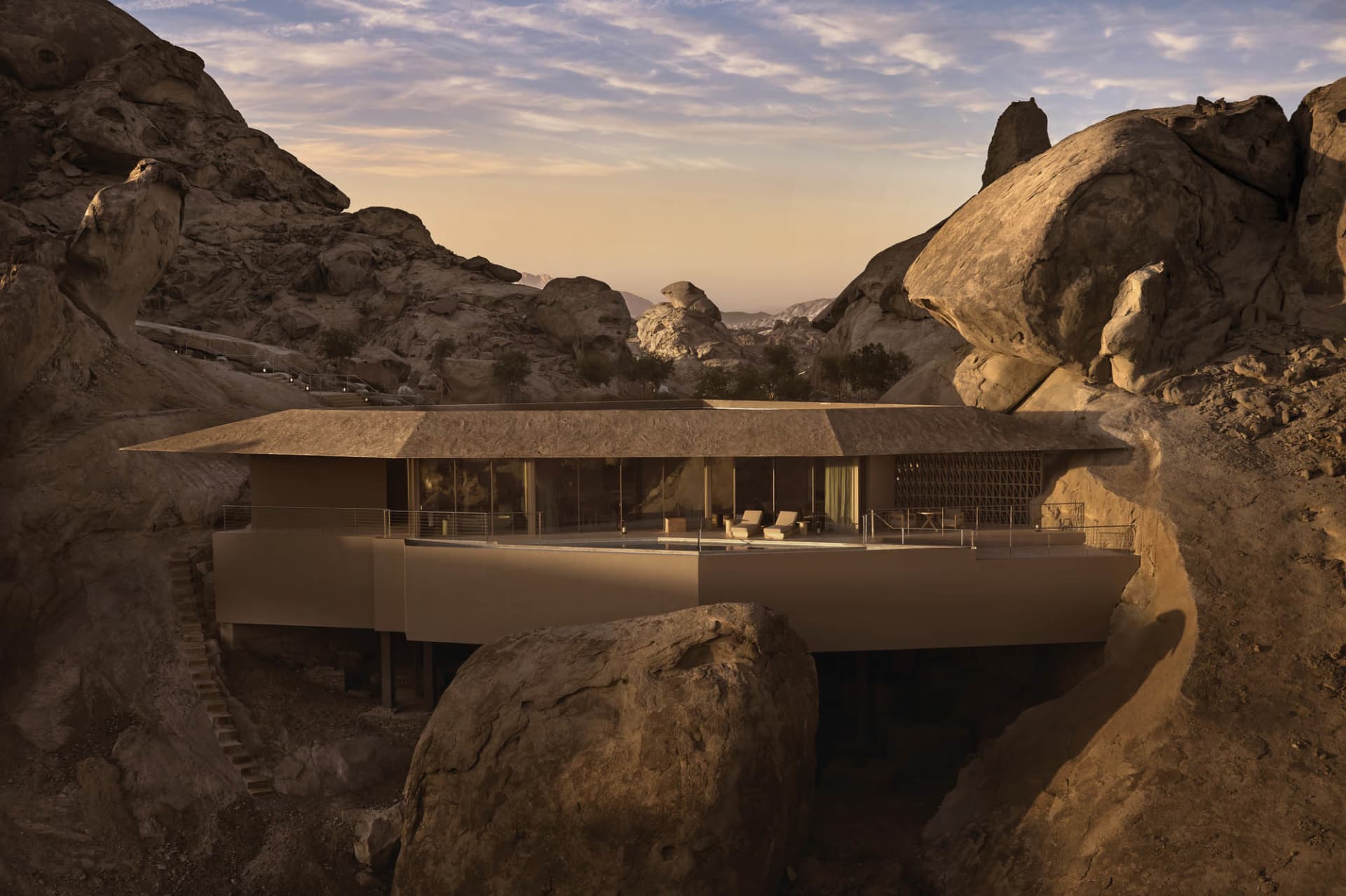 A modern house with a glass facade and concrete construction, situated in front of a large rock in a mountainous landscape, with a bright and inviting interior