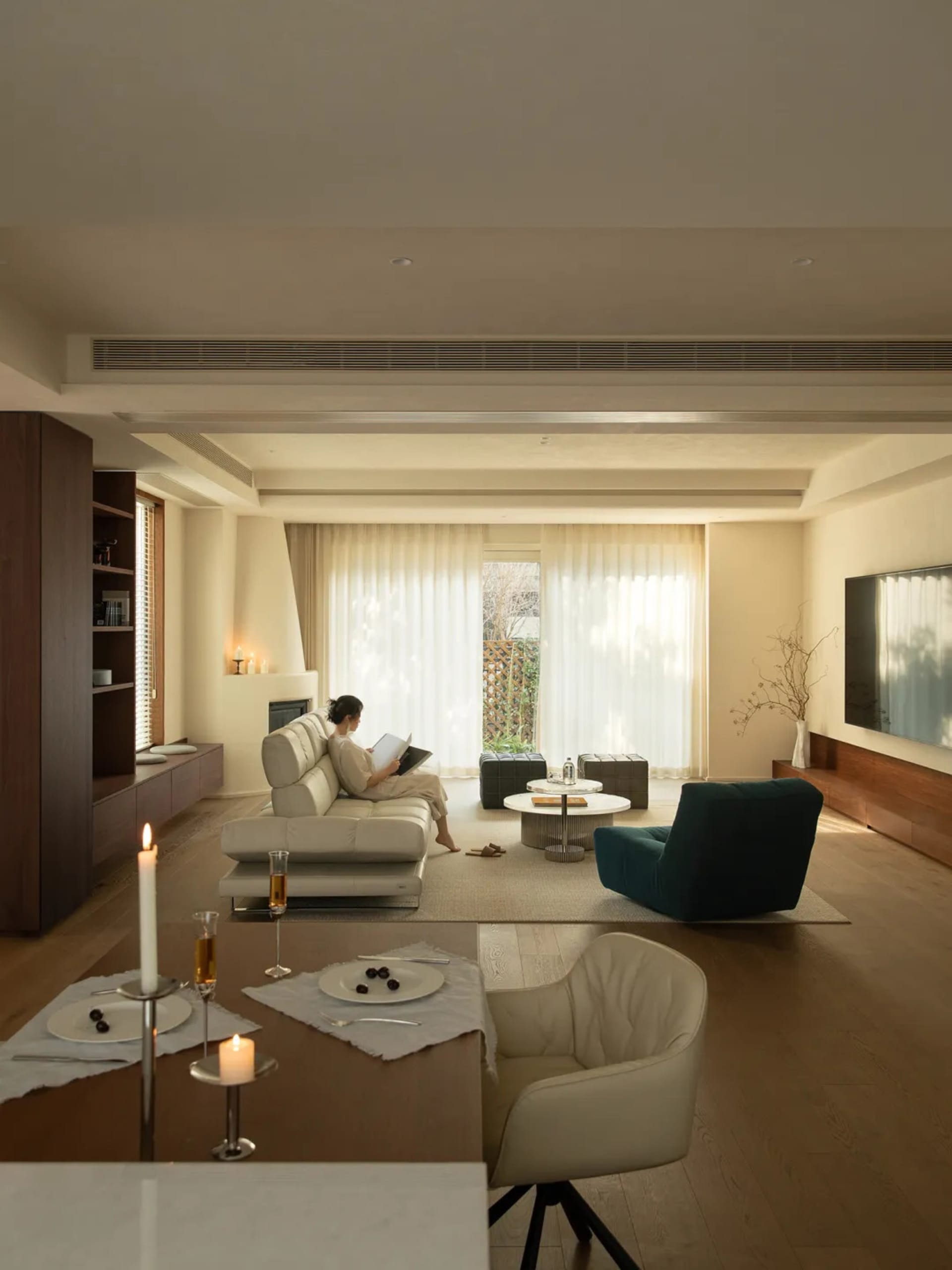 A well-lit living room with a woman on a couch, a dining table, chairs, and a TV, surrounded by white furniture and natural light
