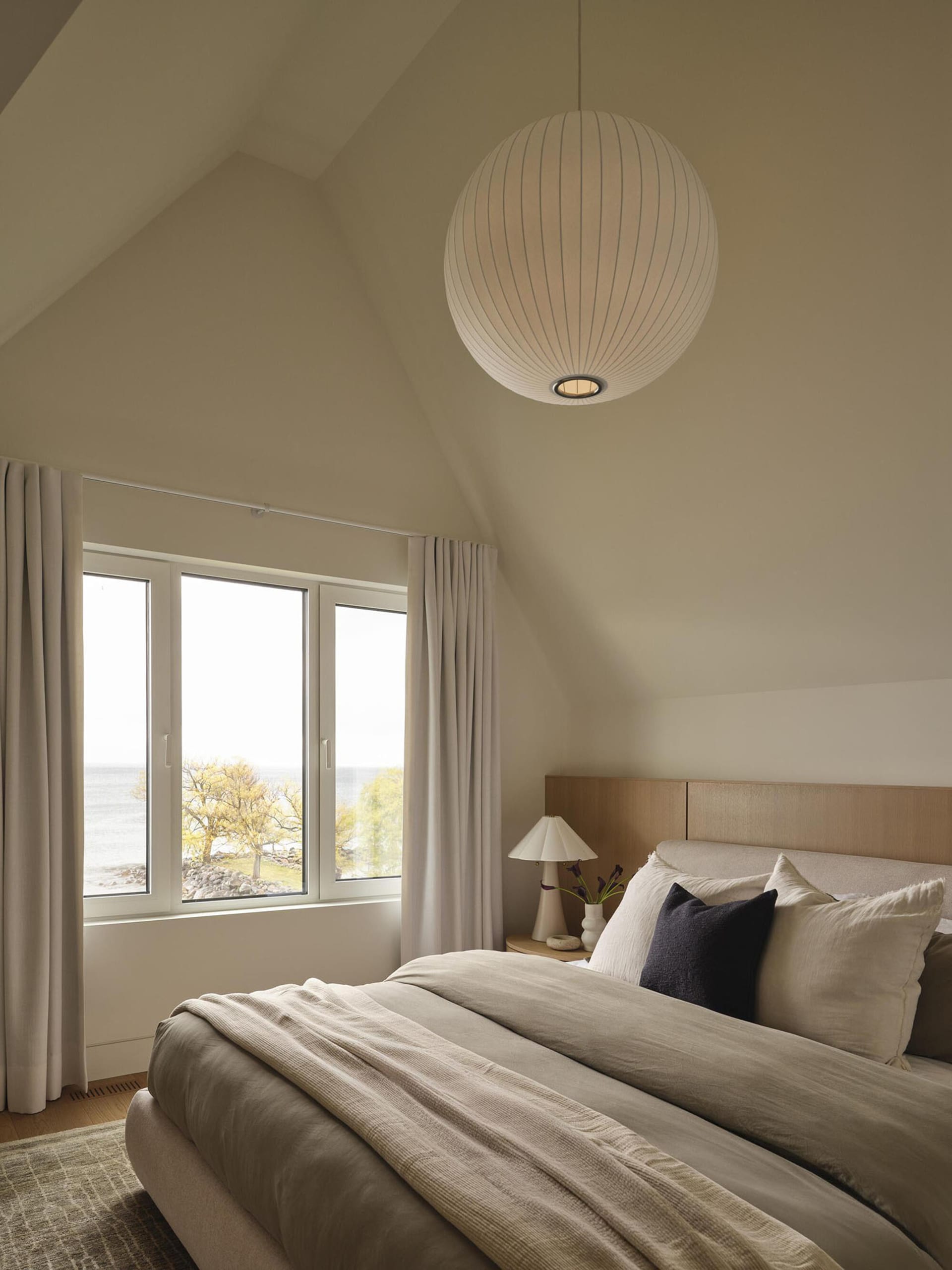 A calm bedroom with a white bed, a window, and a table lamp, surrounded by a white color scheme and natural light