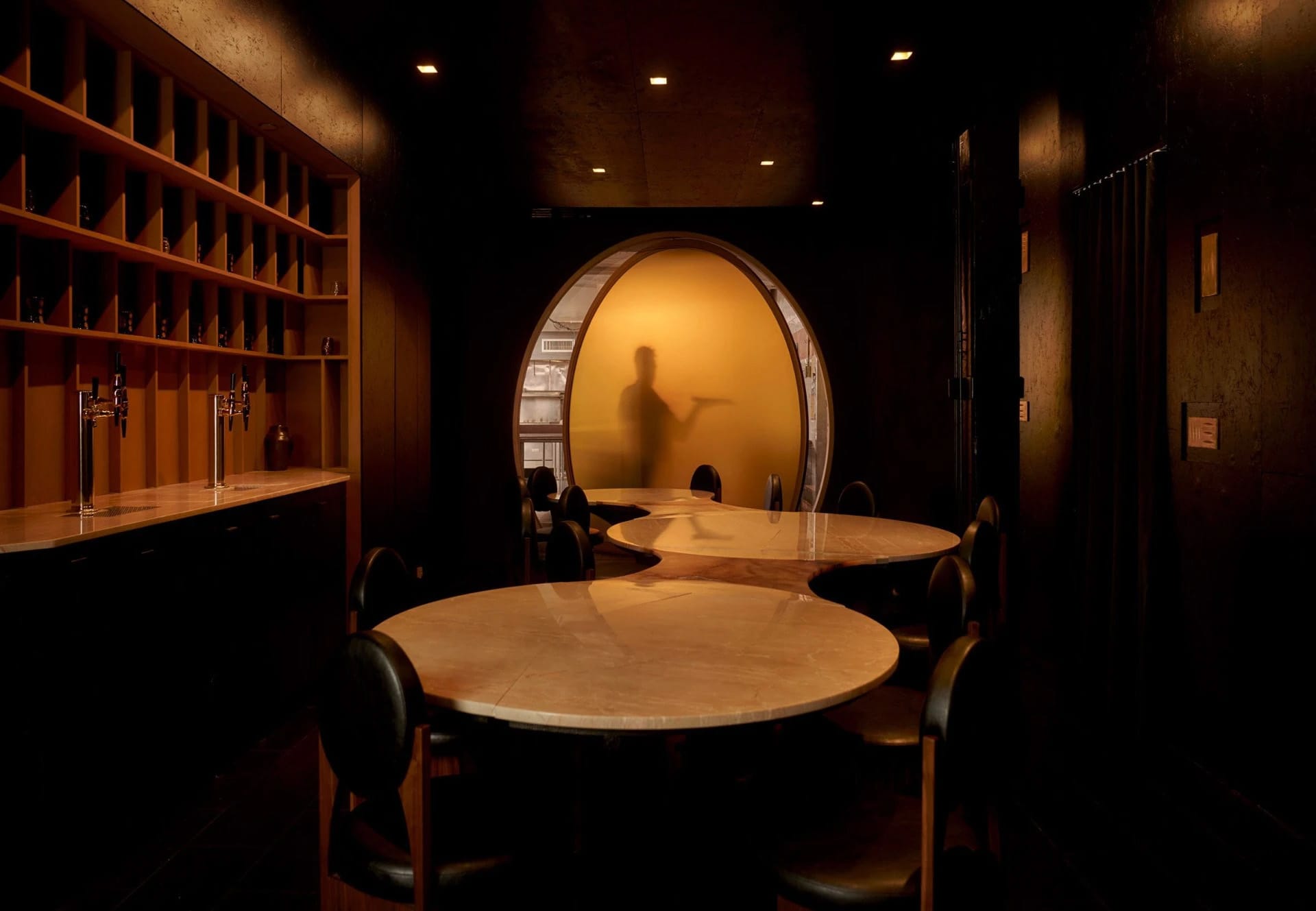 A cozy restaurant dining area with a large round table, wine glasses, and a server, surrounded by wood and glass elements, with soft and warm lighting