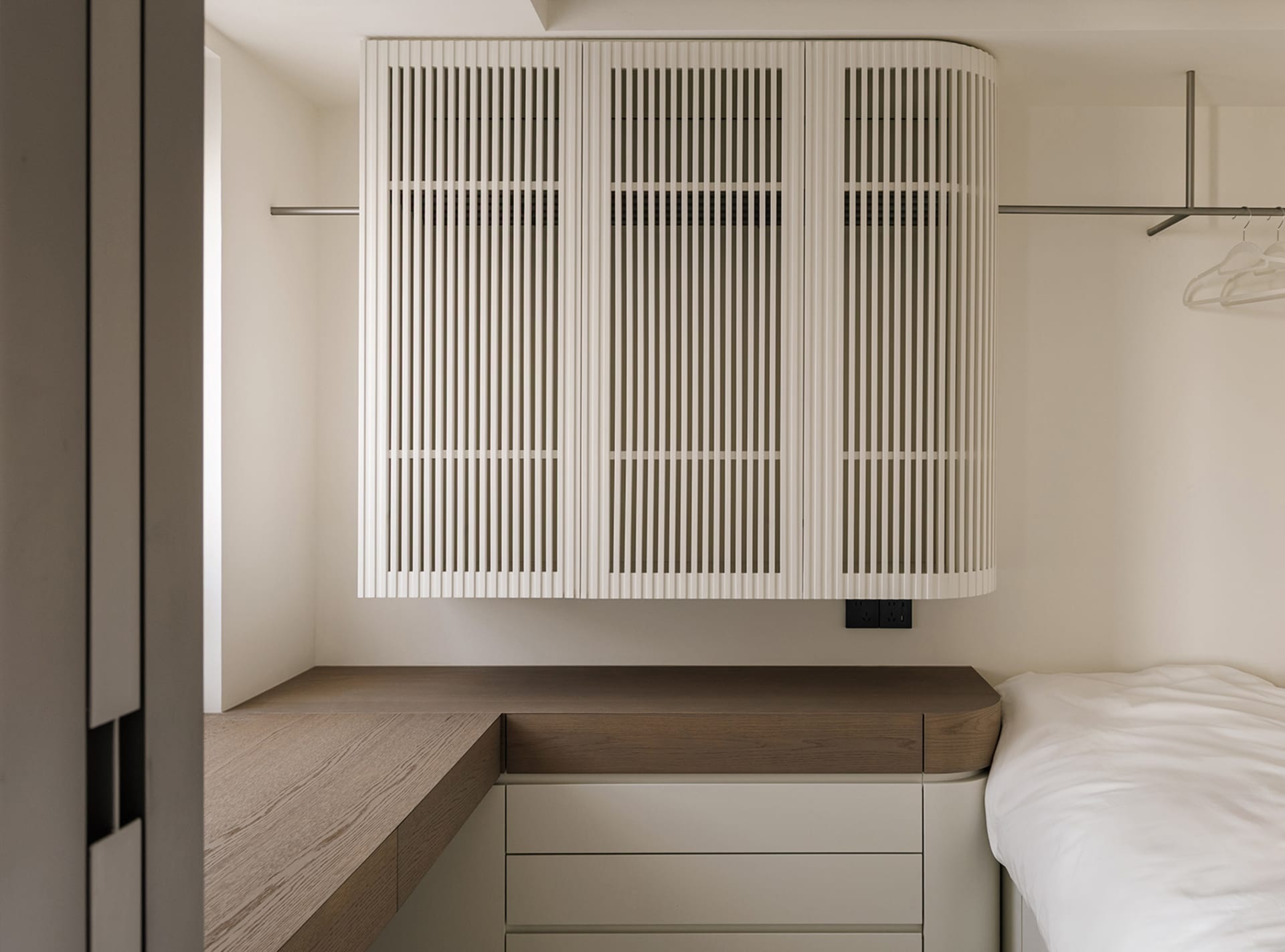 A minimalist bedroom with a wooden desk, white cabinet, and a bed with a white comforter and pillows, lit by natural light