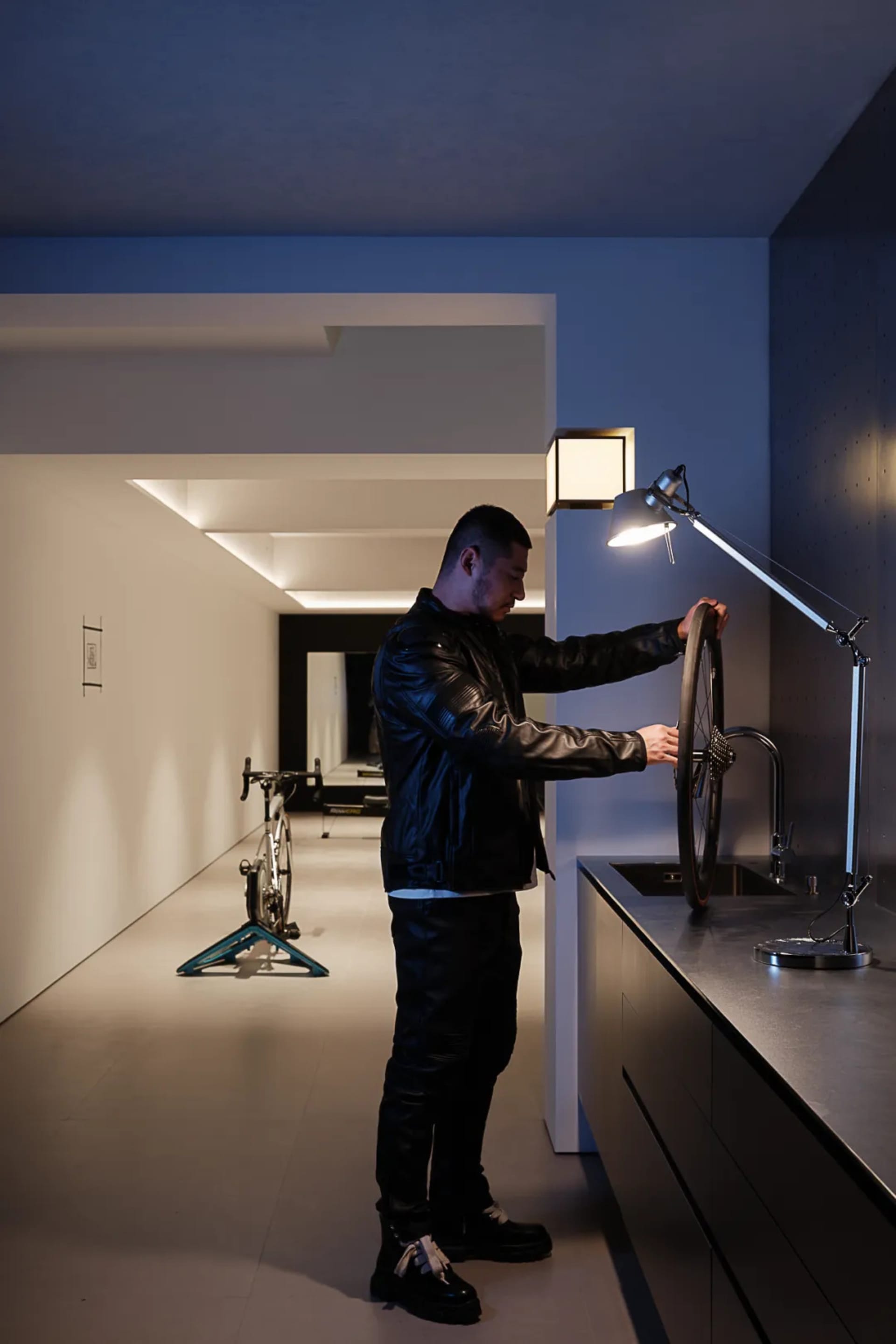 A man in a bathroom, standing next to a sink and a bicycle, with a toilet in the background, wearing a leather jacket and adjusting a bike wheel, in a white-colored room with a light on the wall