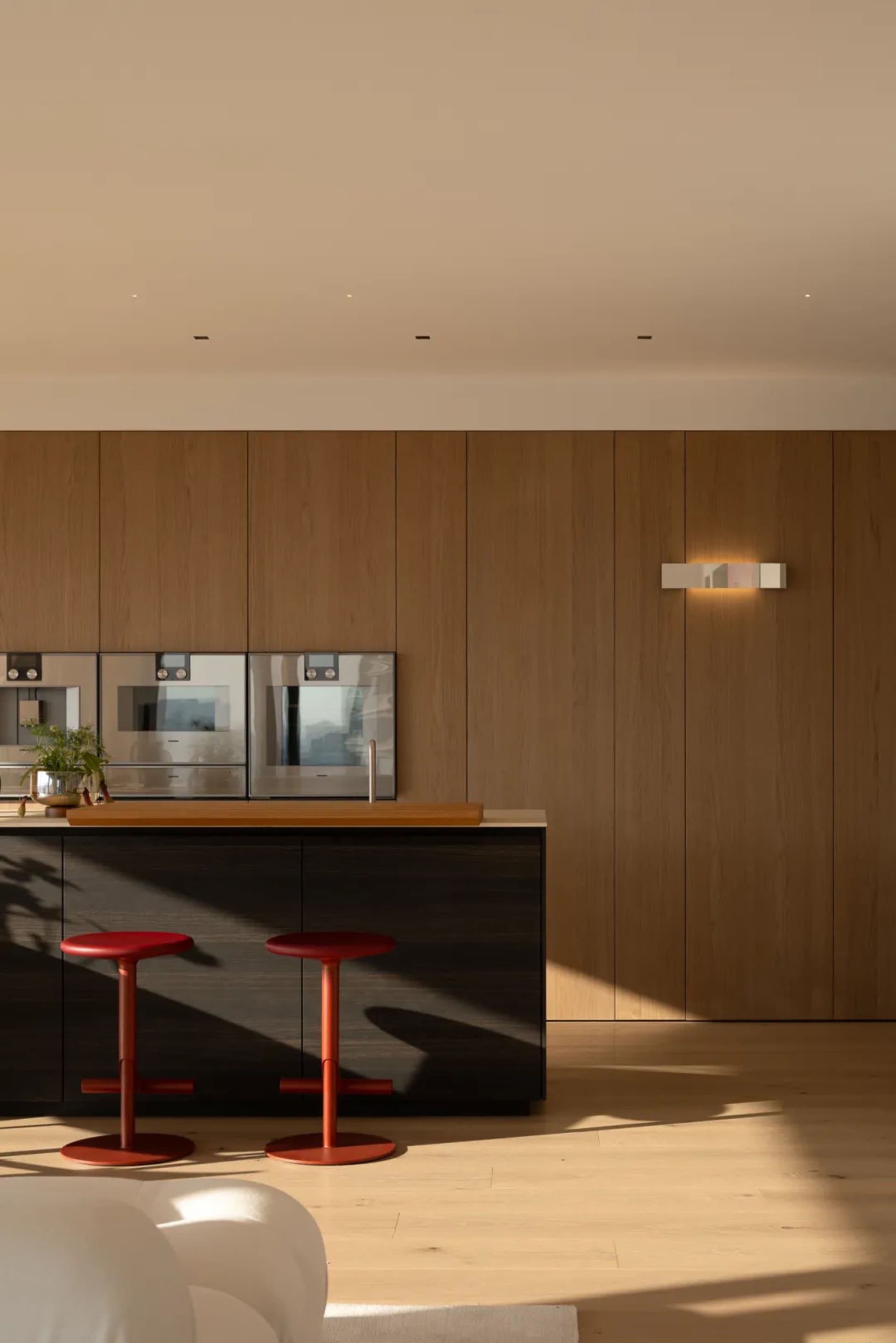 Modern kitchen with wooden walls and cabinets, featuring a counter with a sink, refrigerator, and oven, with two red stools and a potted plant, lit by natural sunlight