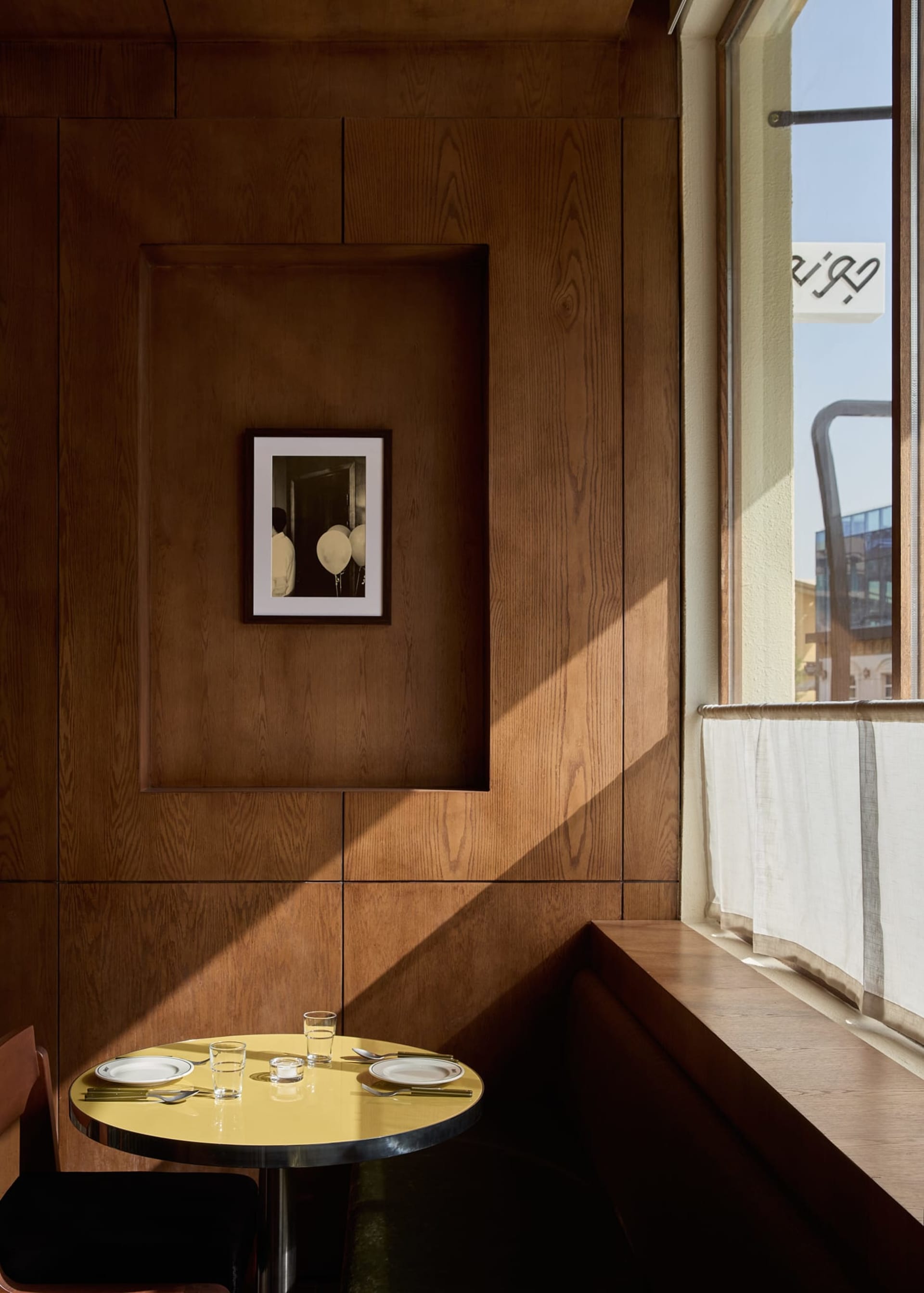A modern dining space with a wooden table, chairs, and bench, set with plates, cups, and a wine glass, surrounded by a wooden wall and natural light